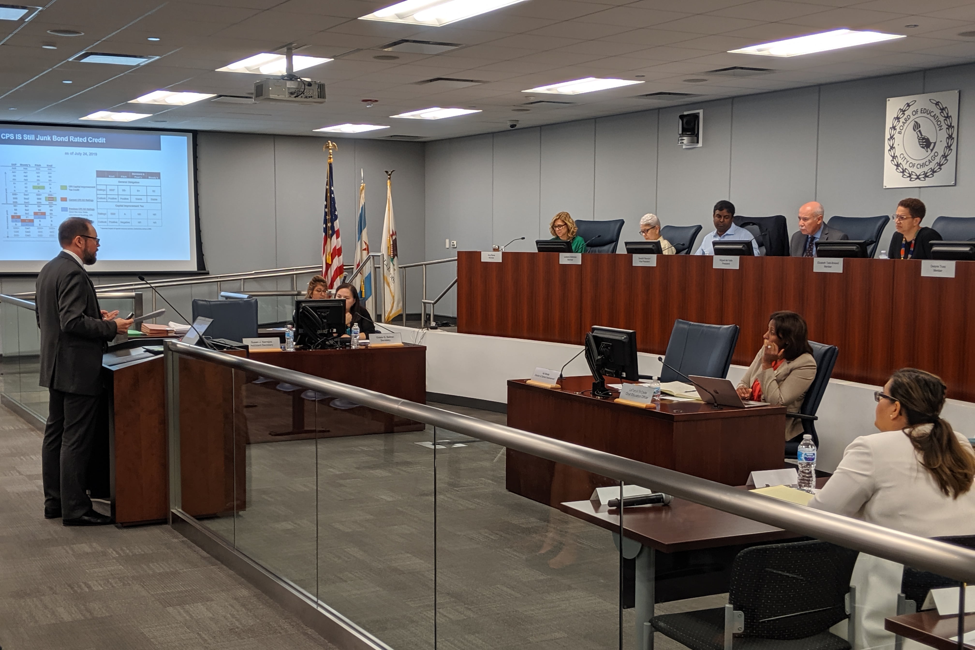 Members of the Chicago Board of Education sit behind a brown desk as they listen to a person in a black suit giving public comment during their monthly board meeting.