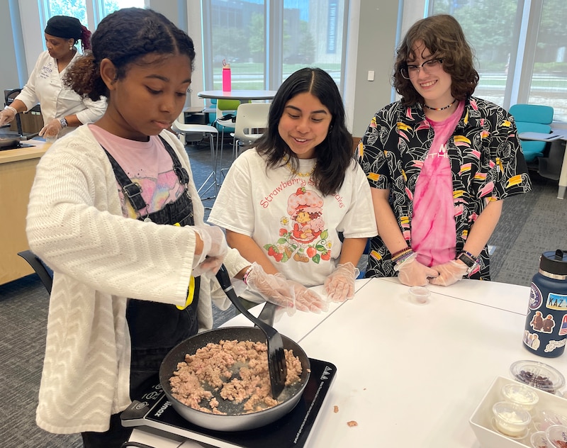 A student with a white sweater stirs food in a pan while two other students watch.