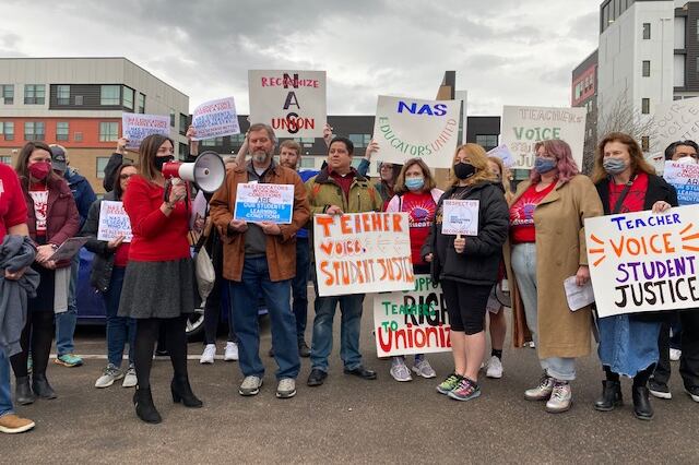 A group of people wearing masks and coats and holding signs that read “Teacher Voice Student Justice” cluster together. On the left side, a woman in a red blazer and dark skirt holds a bullhorn and addresses the crowd. Mid-rise apartment buildings can be seen behind them.