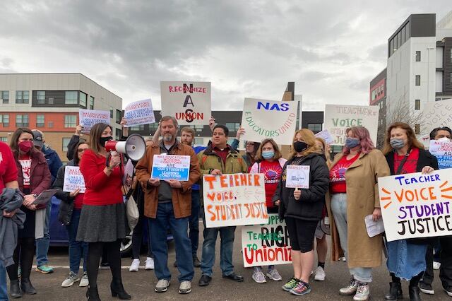 A group of people wearing masks and coats and holding signs that read “Teacher Voice Student Justice” cluster together. On the left side, a woman in a red blazer and dark skirt holds a bullhorn and addresses the crowd. Mid-rise apartment buildings can be seen behind them.