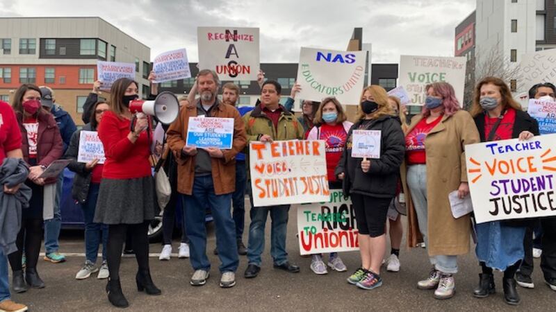 A group of people wearing masks and coats and holding signs that read “Teacher Voice Student Justice” cluster together. On the left side, a woman in a red blazer and dark skirt holds a bullhorn and addresses the crowd. Mid-rise apartment buildings can be seen behind them.