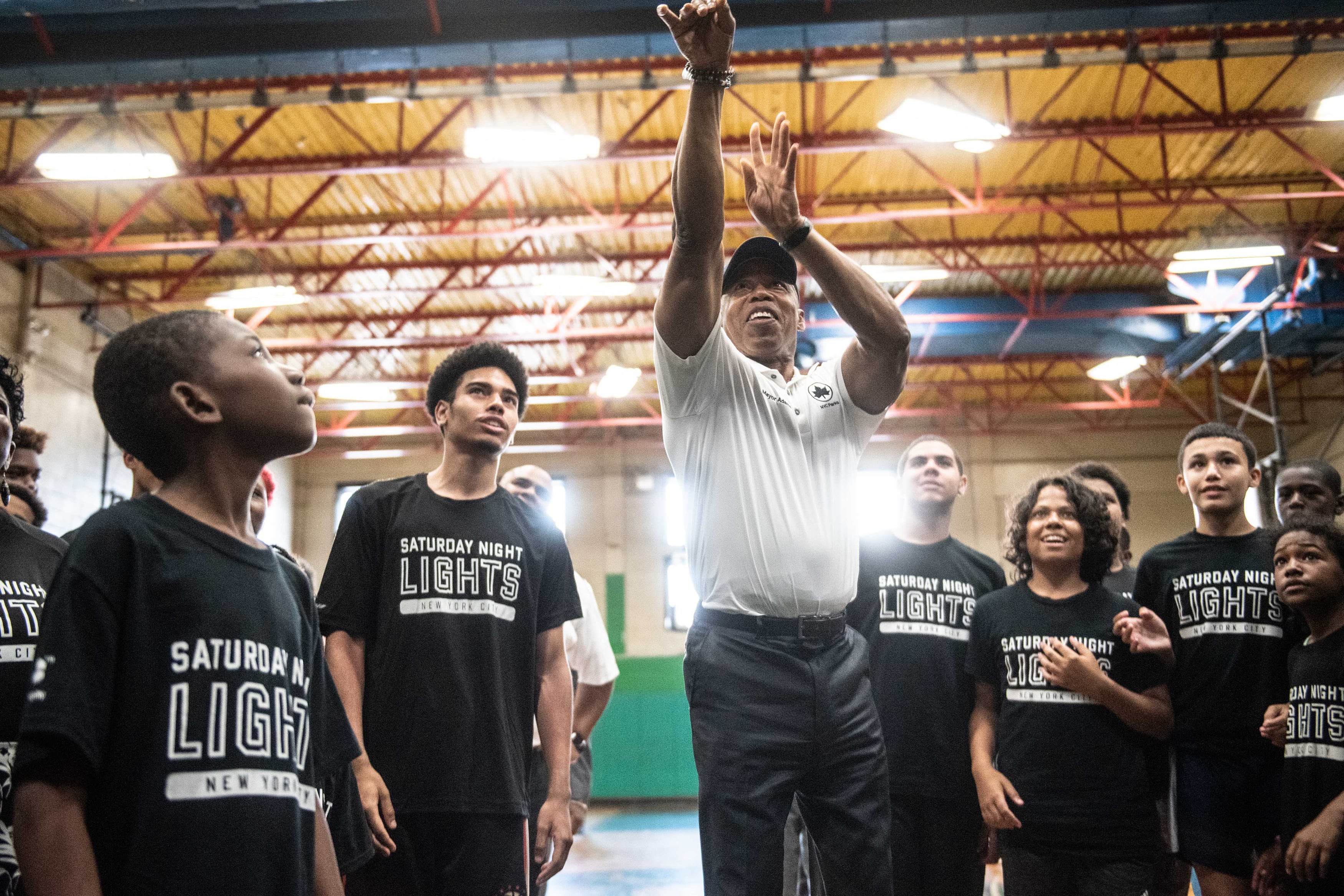 A man wearing a white dress shirt shoots a basketball with a group of students wearing the same shirts stands around.