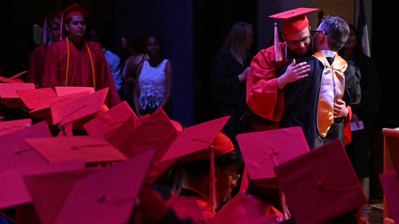 A high school graduate in a red cap and gown is hugged in front of a sea of other graduates