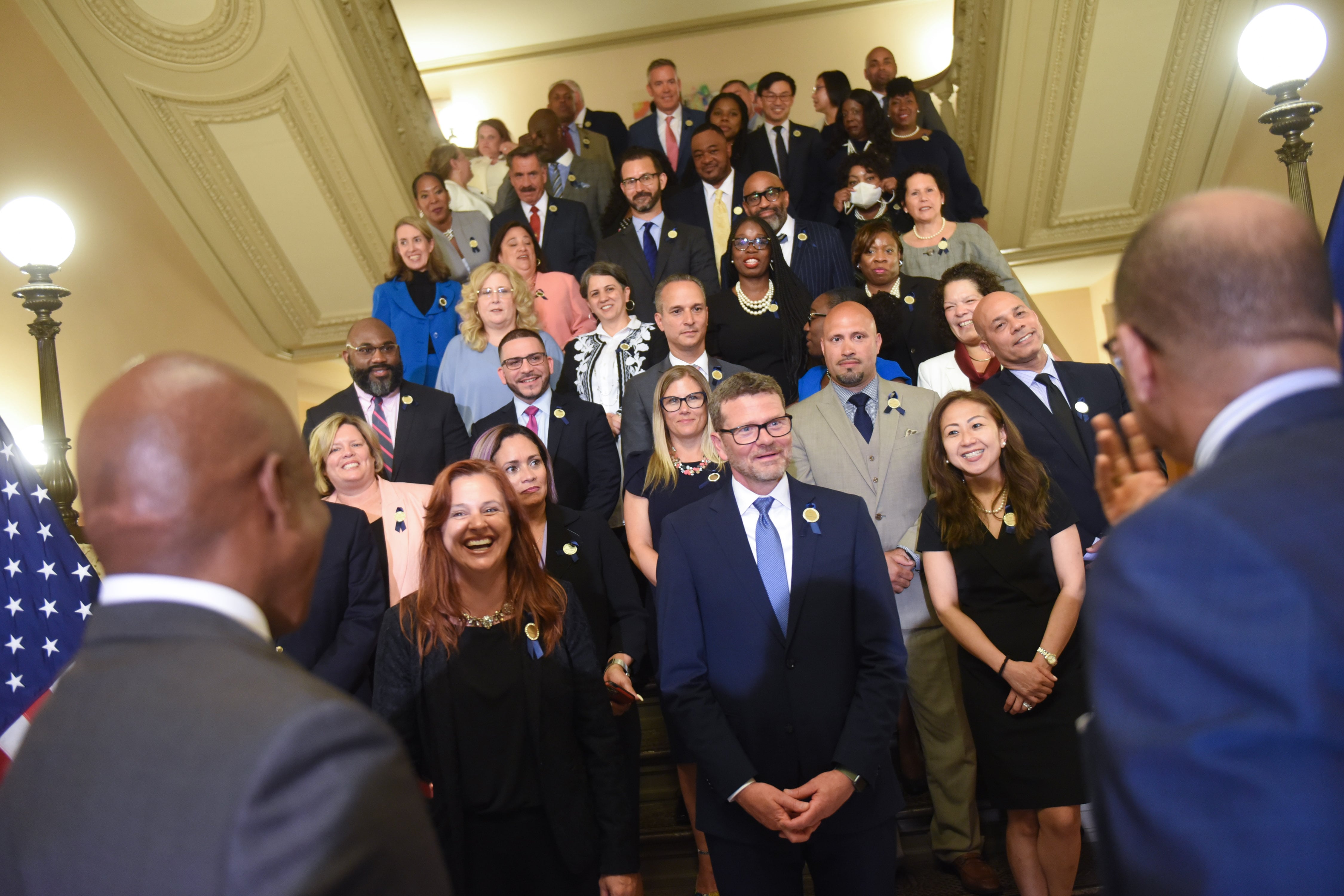 Two men face away from the camera and toward a group of smiling people on stairs
