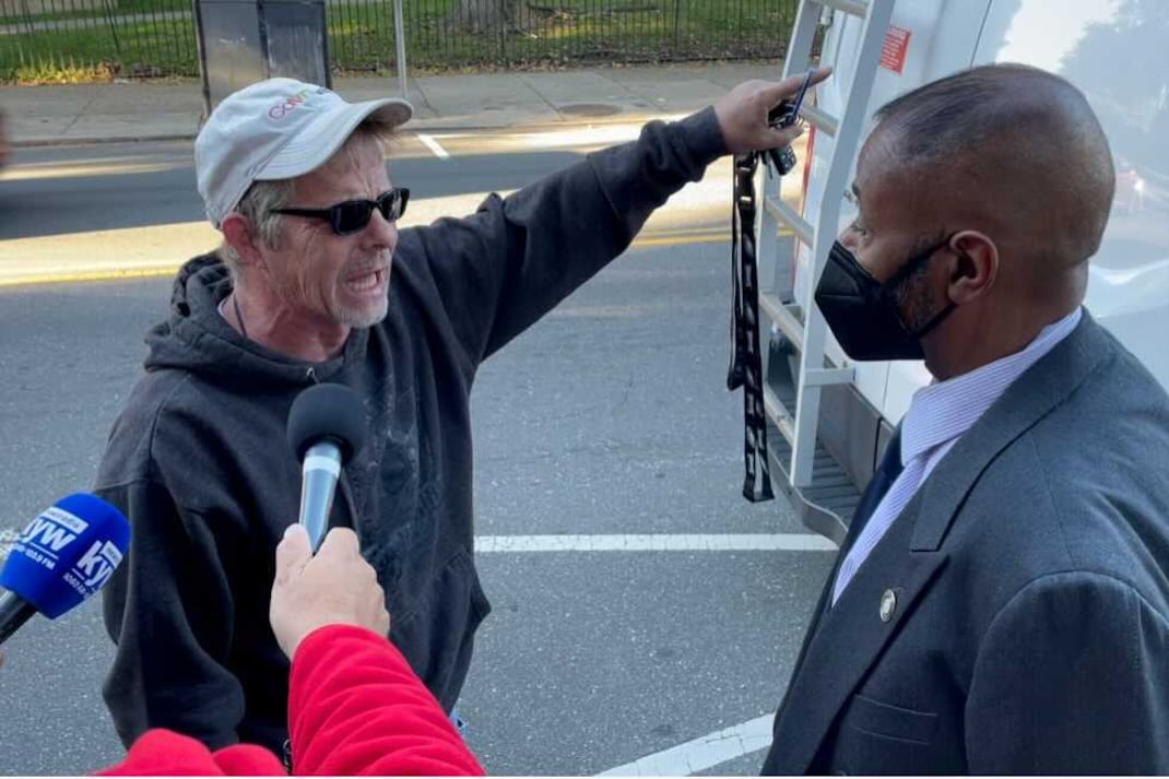 A man in a white hat wearing sunglasses speaks to a man wearing a suit, collared shirt, and a mask, while another person holds a microphone.