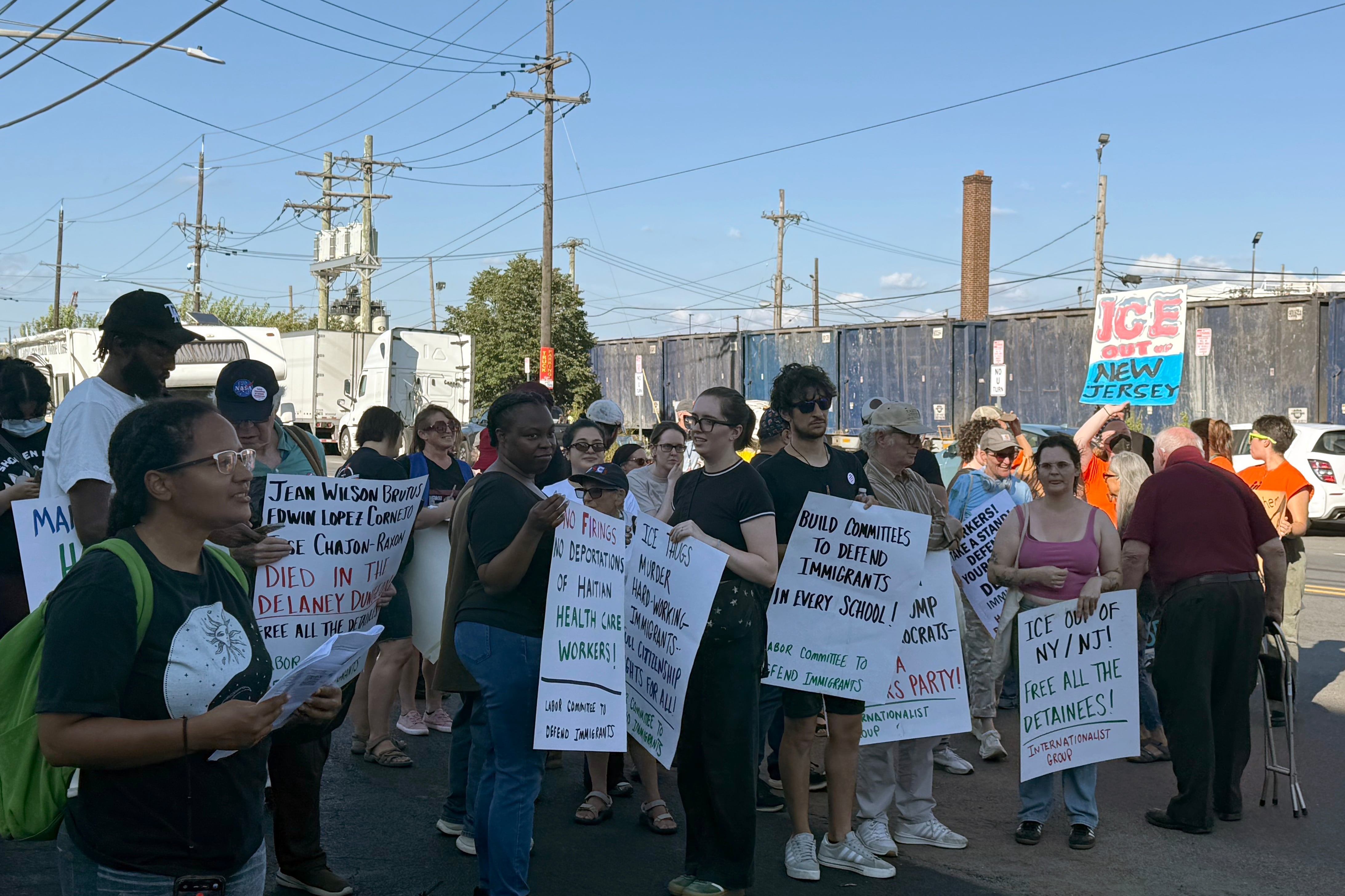 A group of adults, some holding signs, stand outside during a rally against ICE and deportations.