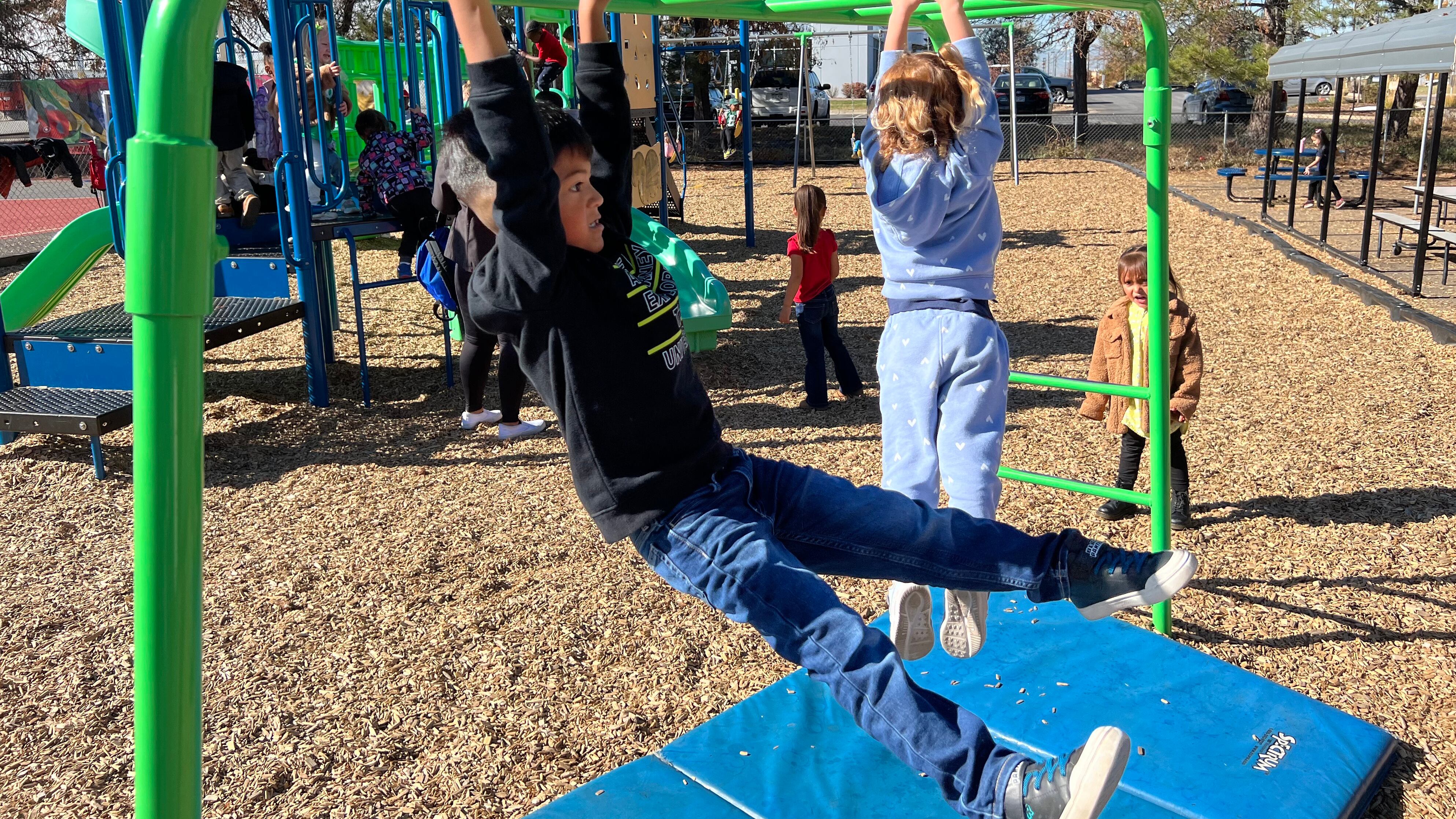 Two young kinds swing on bars at a playground outside with other young students in the background.