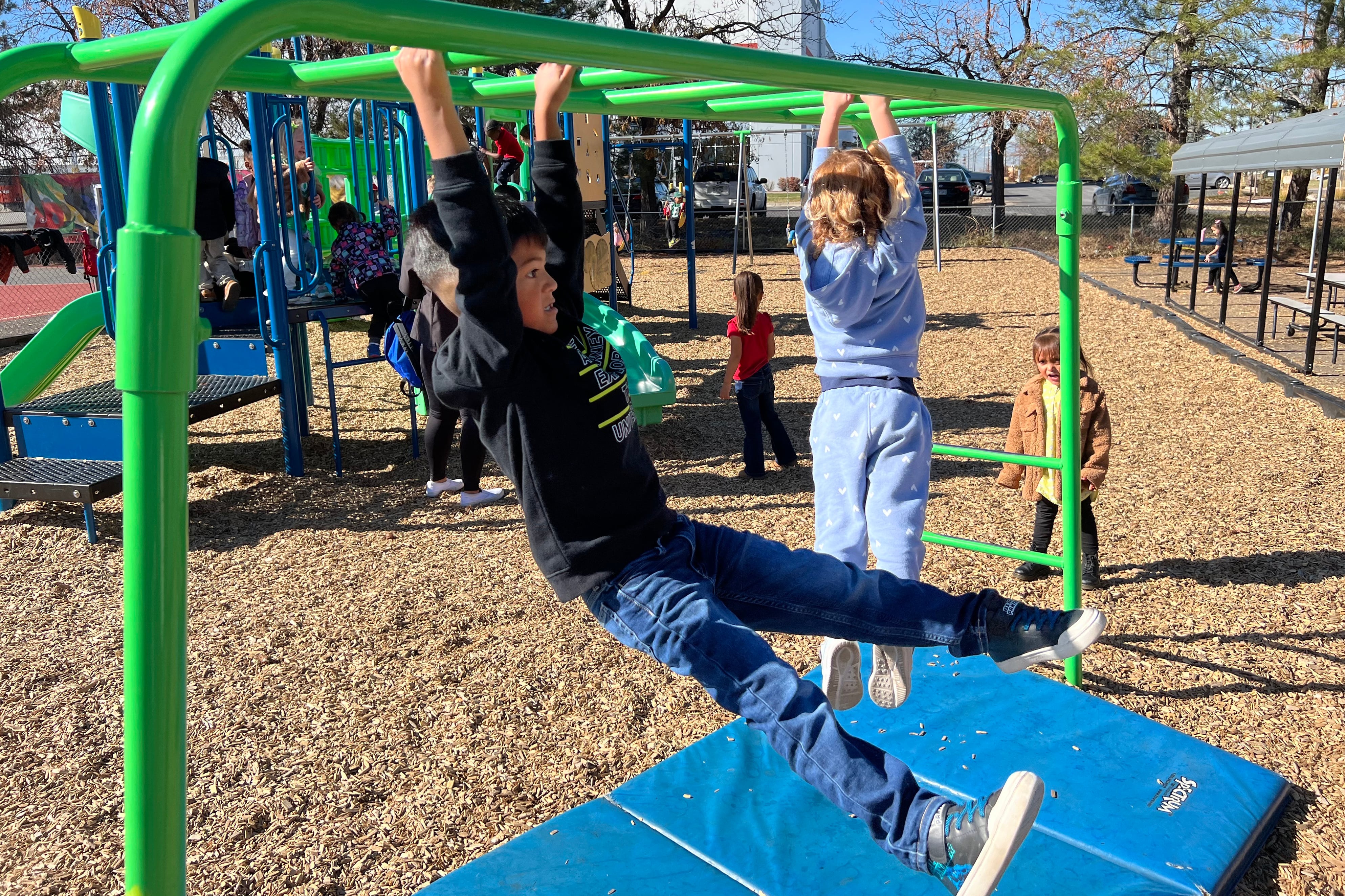 Two young kinds swing on bars at a playground outside with other young students in the background.