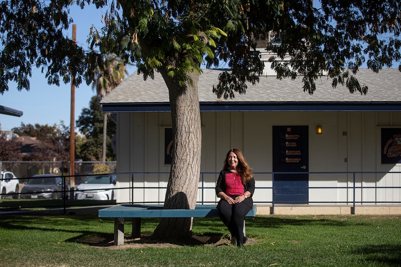 A woman in a red top and black pants sits on a bench under a tree.