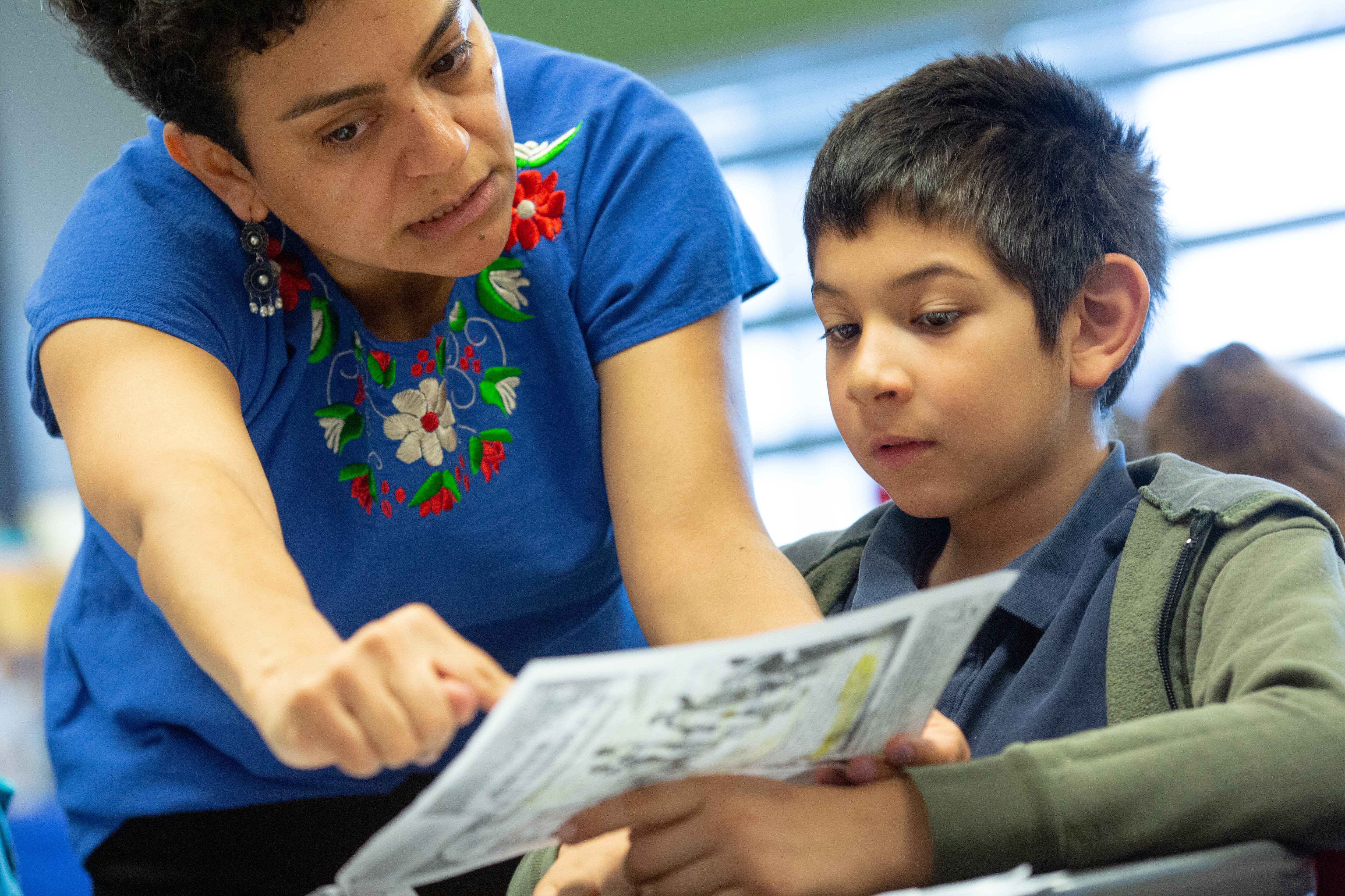 An elementary school teacher leans over a student at his desk while helping him with a writing activity.