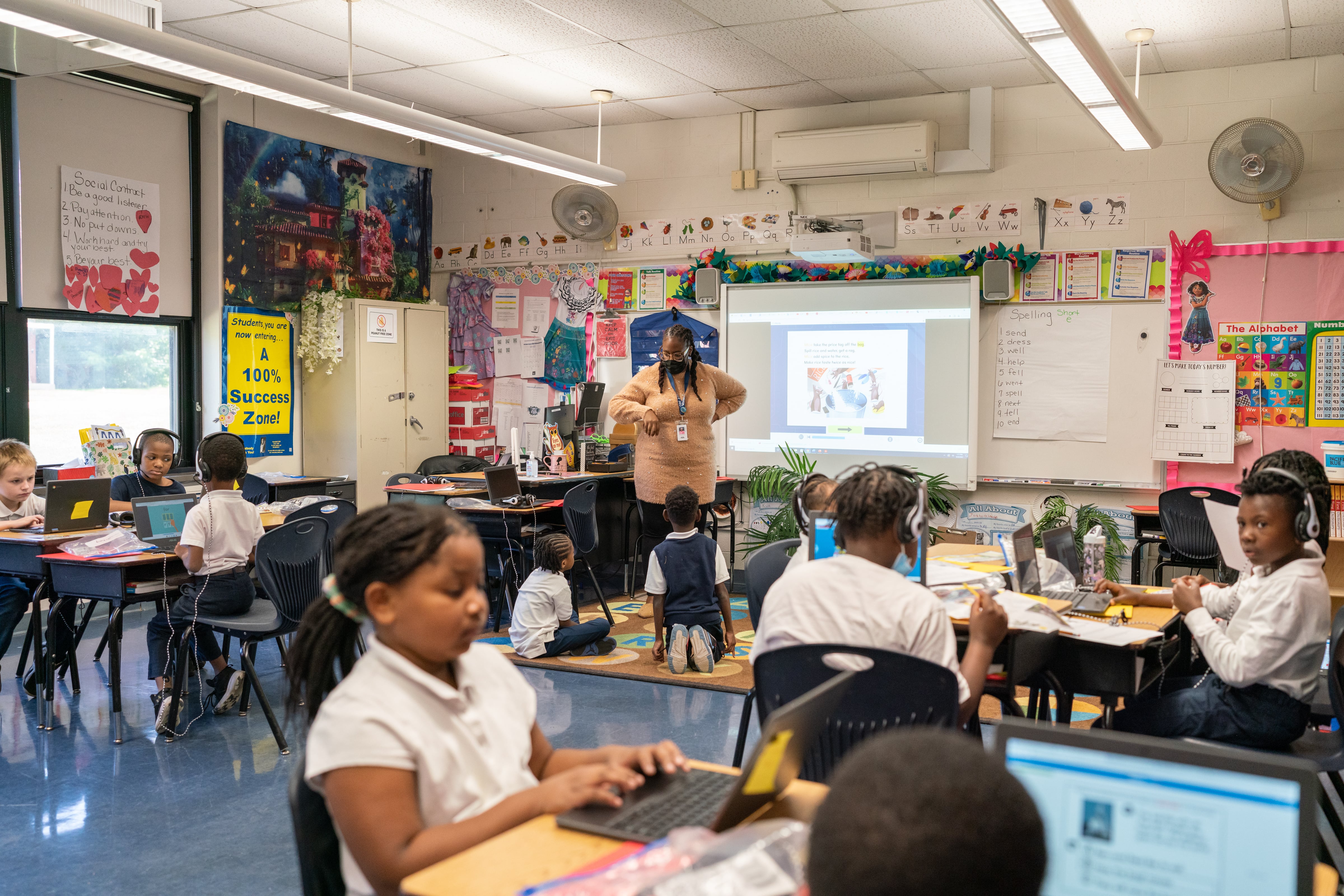 Students work on their laptops in small grouped desks in a classroom. Their teacher leads the class and stands next to a projector in the front of the room.