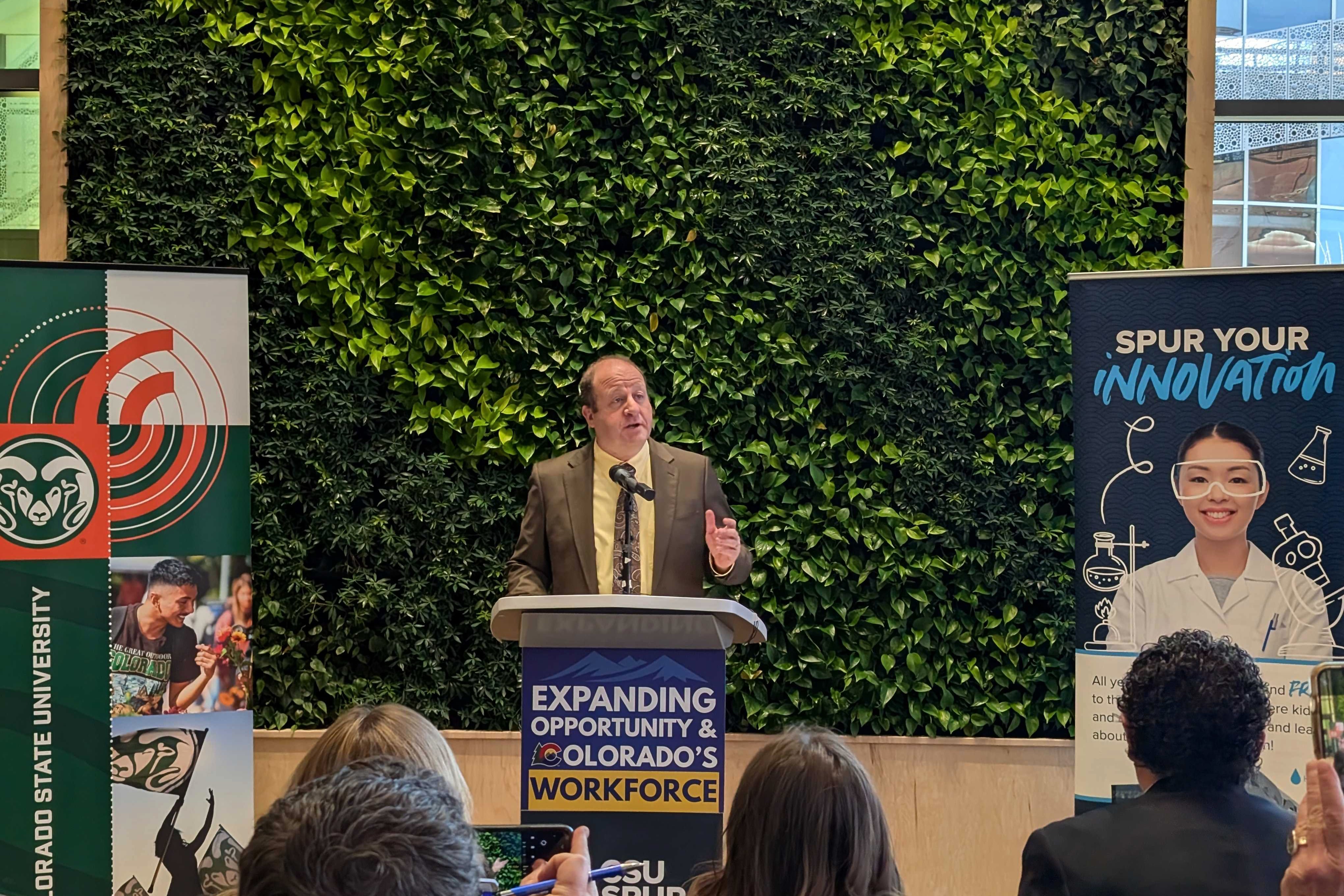 A photograph of a white man in a suit speaking from a stage with greenery in the background and the backs of peoples' heads sitting in the crowd in the foreground.