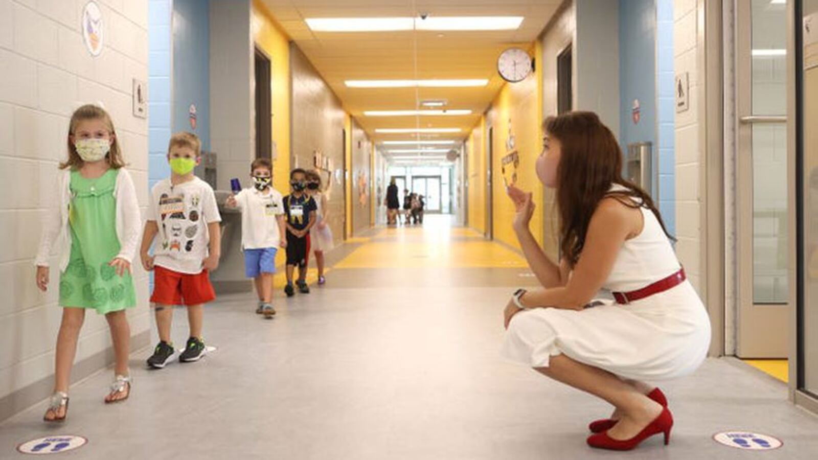 Woman in white sleeveless shift with red patent belt and shoes squats in school hallway, waving at a line of elementary children walking past.