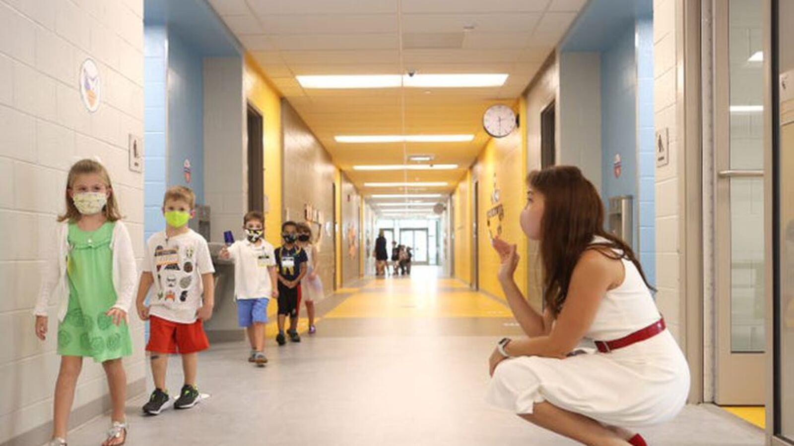Woman in white sleeveless shift with red patent belt and shoes squats in school hallway, waving at a line of elementary children walking past.