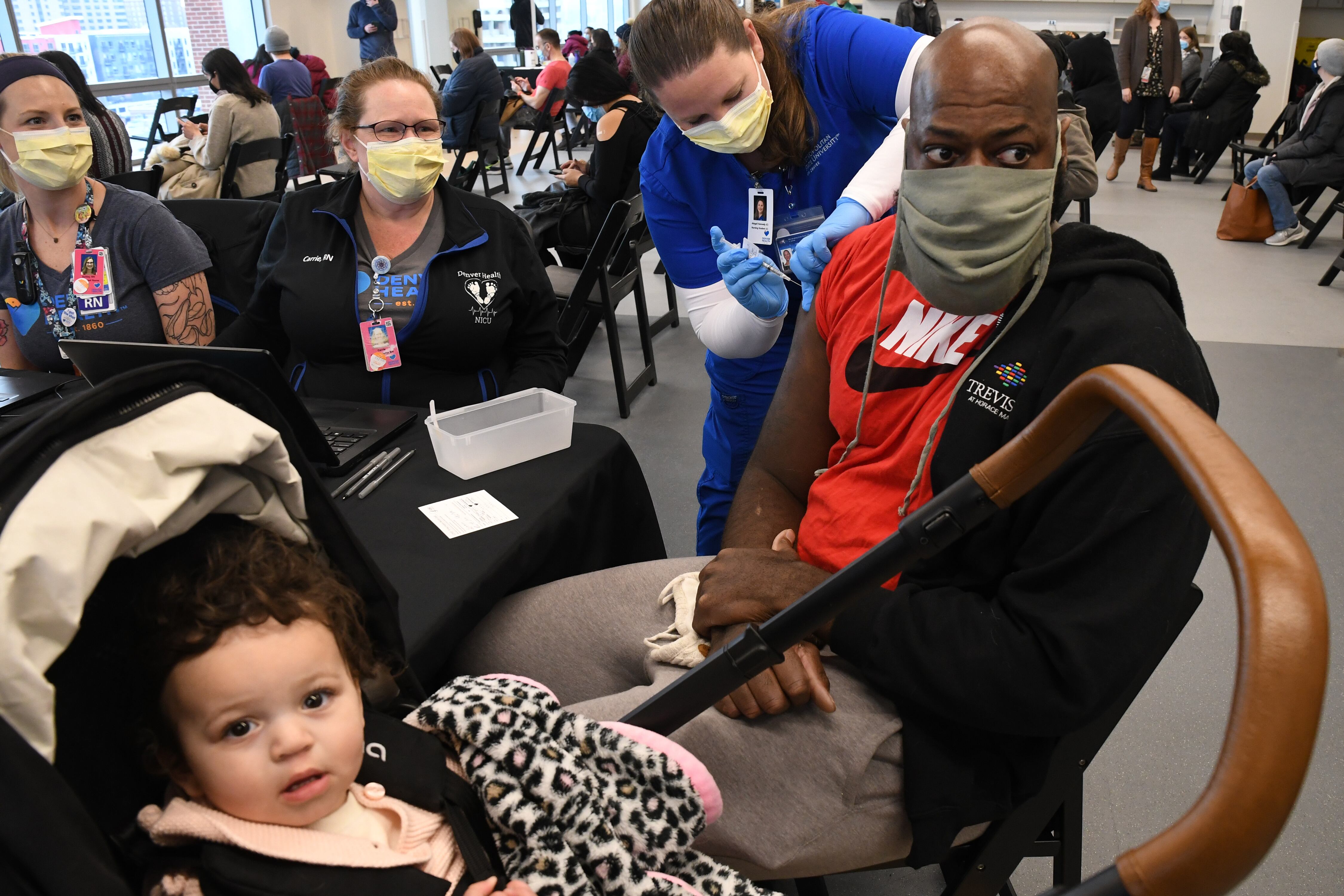 A seated man in a mask gets a COVID vaccine from a masked woman in scrubs. A baby in a stroller looks at the camera.