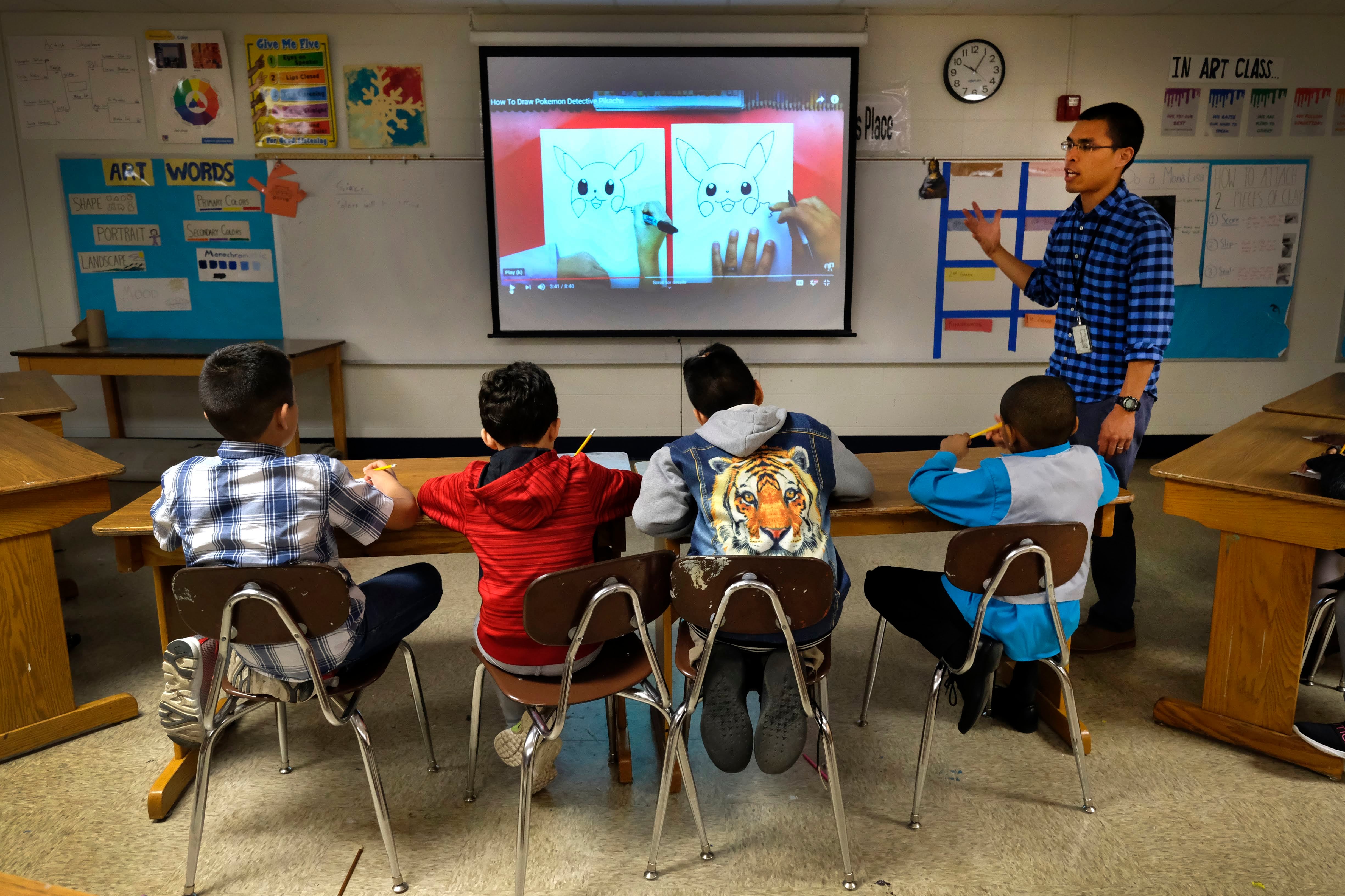 An April 2019 photo shows a teacher next to a smart board talking to four male students sitting at a table and looking at the board.