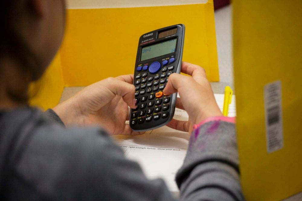 A close up of a student holding a calculator while sitting at a desk in a classroom.