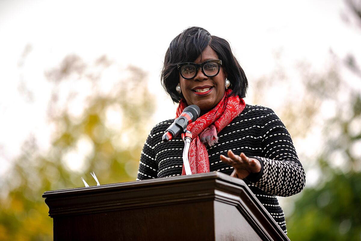 A Black woman in a black shirt with white stripes speaks at a podium