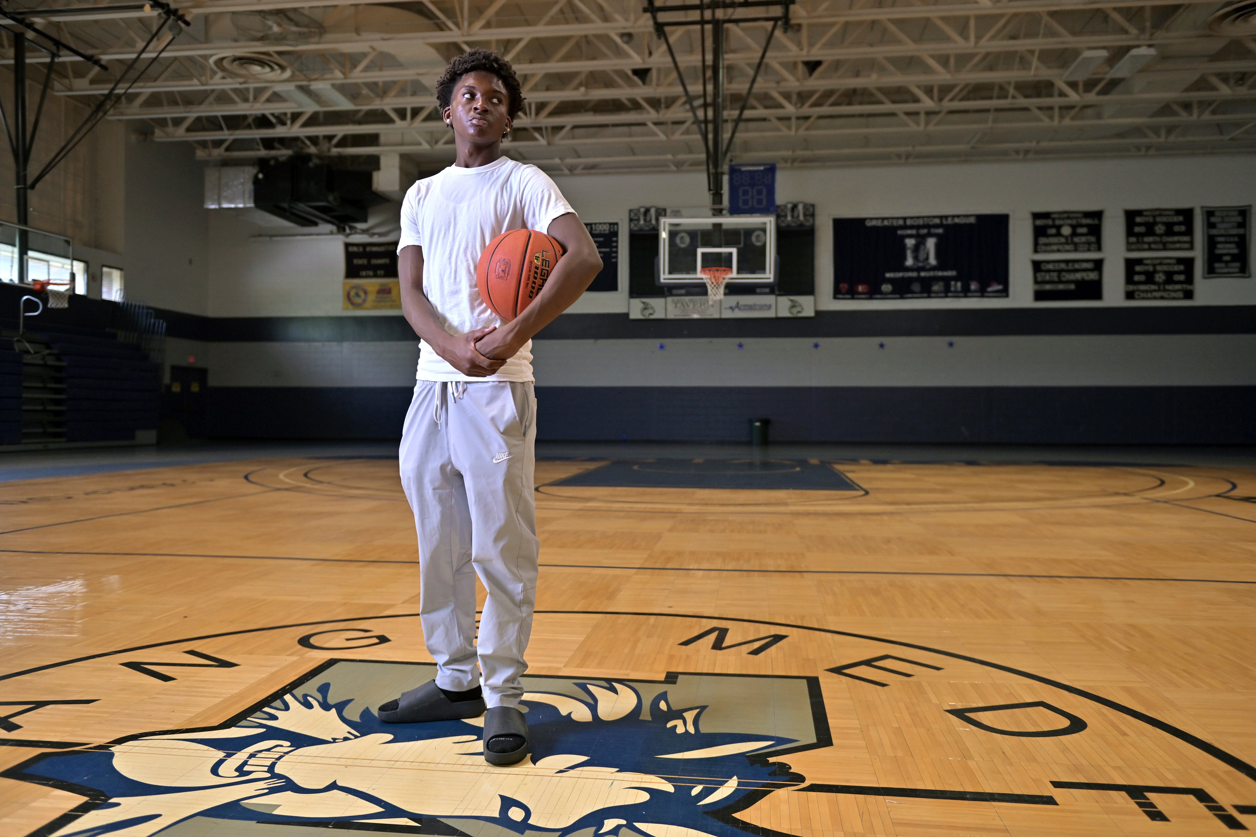 A high school student wearing a white t-shirt, poses for a photo holding a basketball in a gym.