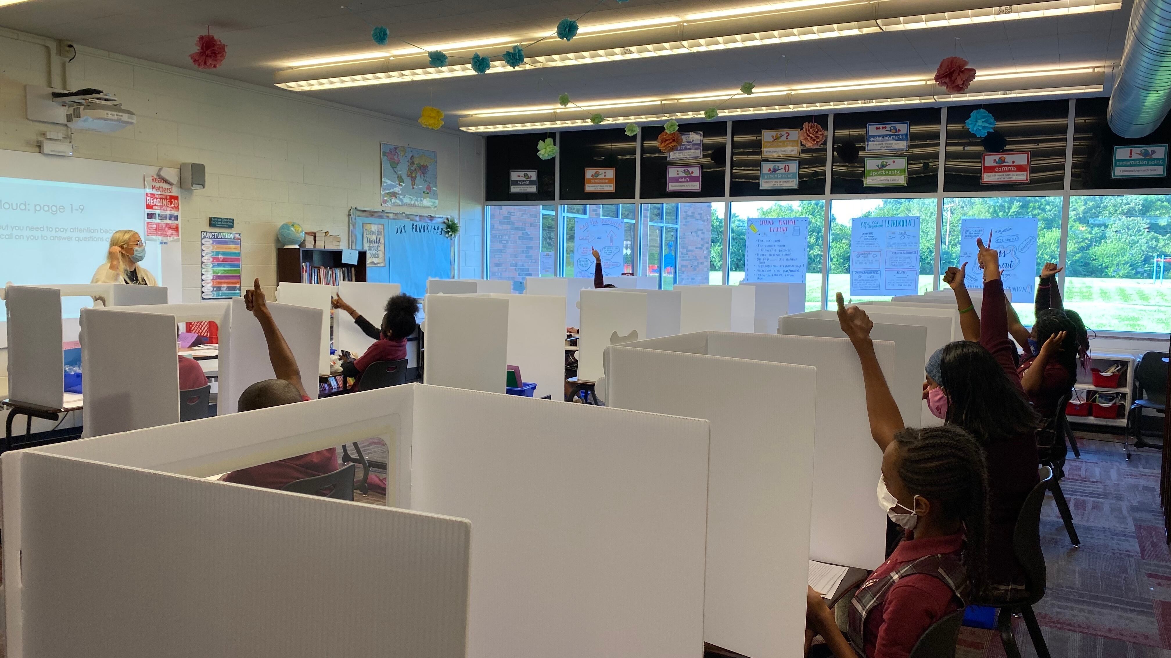 Students separated by partitions raise their hands during class. The teacher stands at the front. 