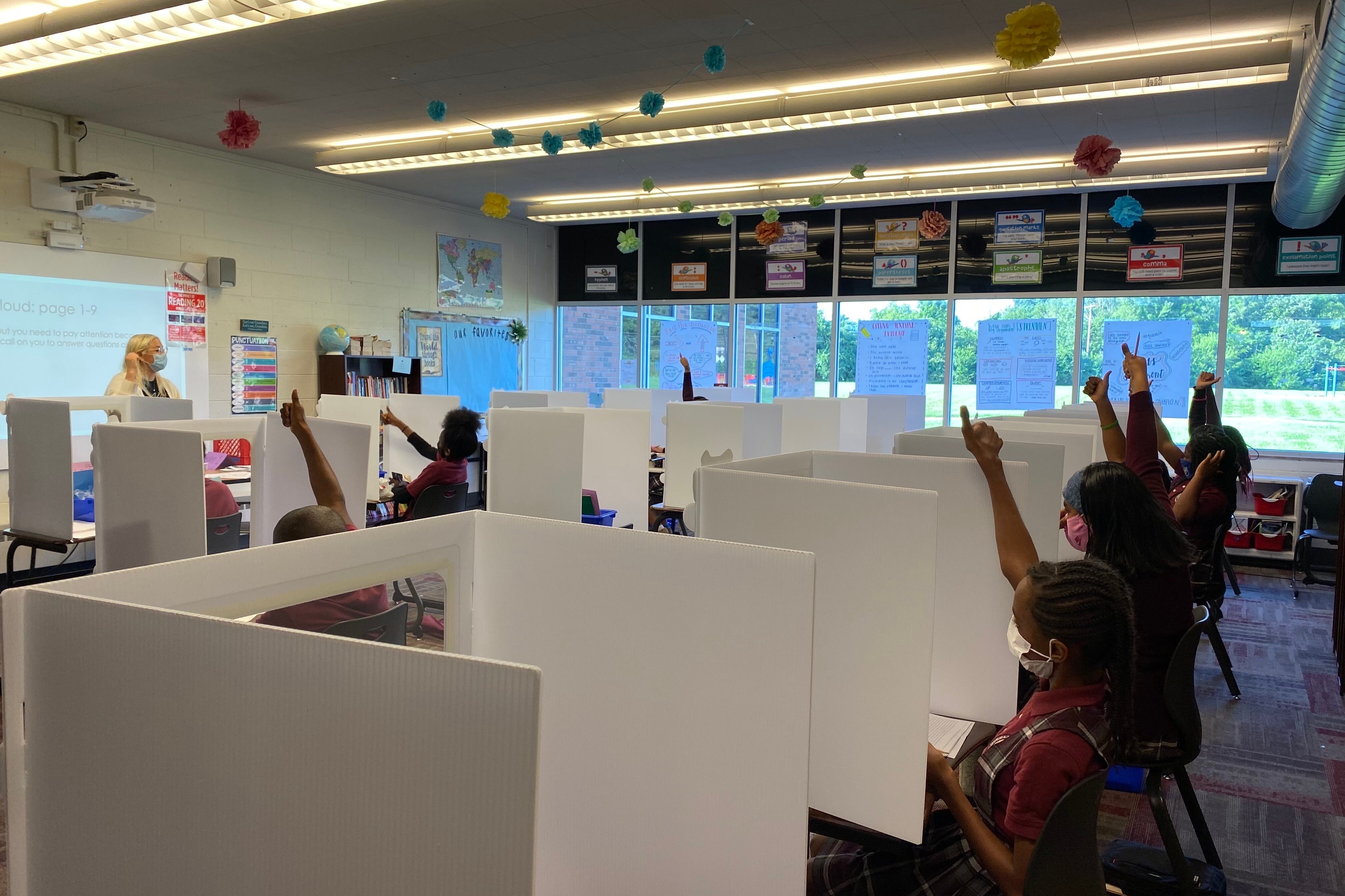 Students separated by partitions raise their hands during class. The teacher stands at the front.