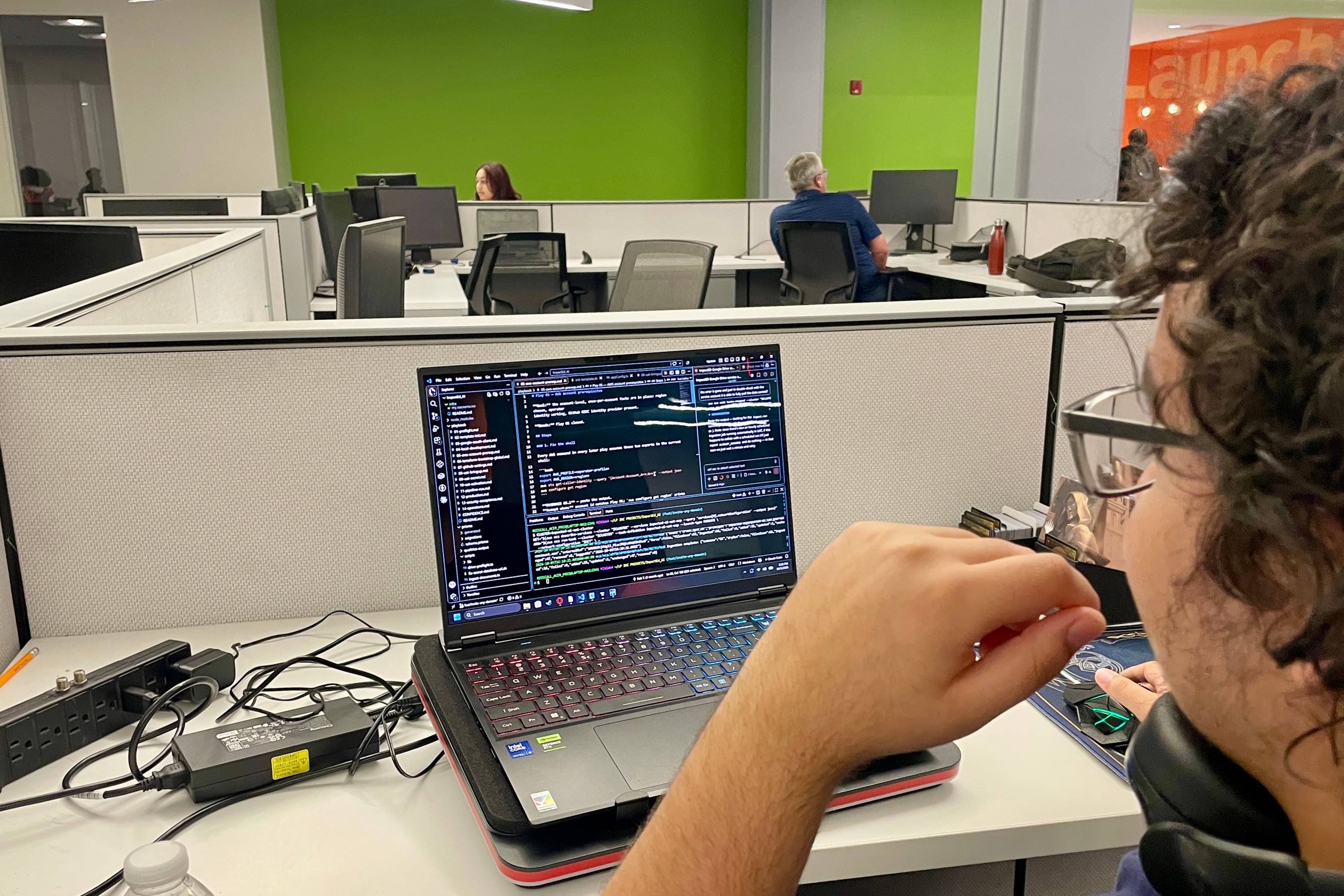 A person works at a laptop at a desk with another person sitting at a computer desk in the background.