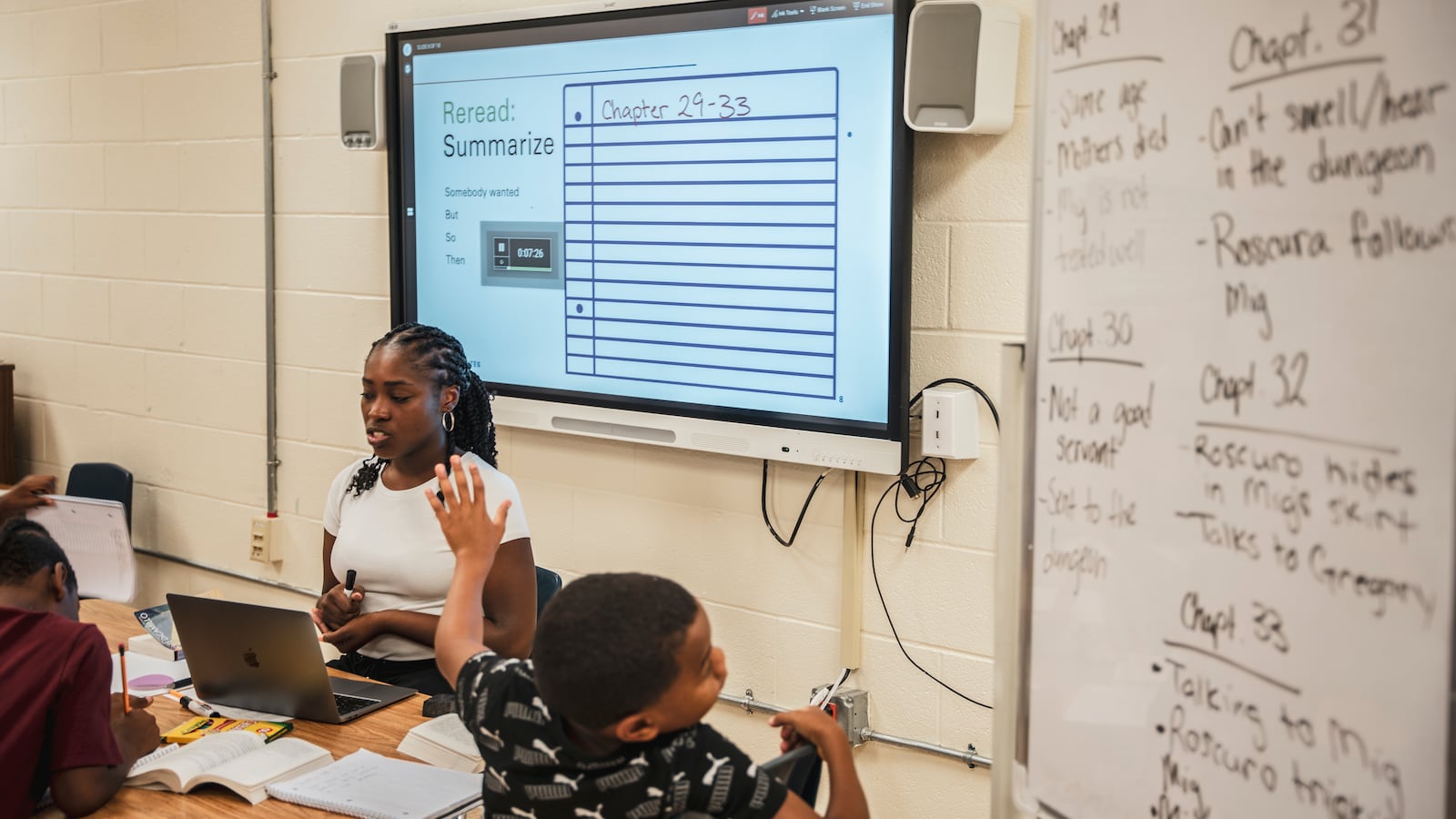 A woman sits at a desk and looks at a laptop as a child raises his hand in front of her.