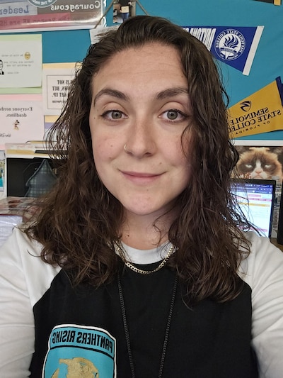 Headshot of a young woman with brown hair. She sits in a classroom and wears a black and white top.