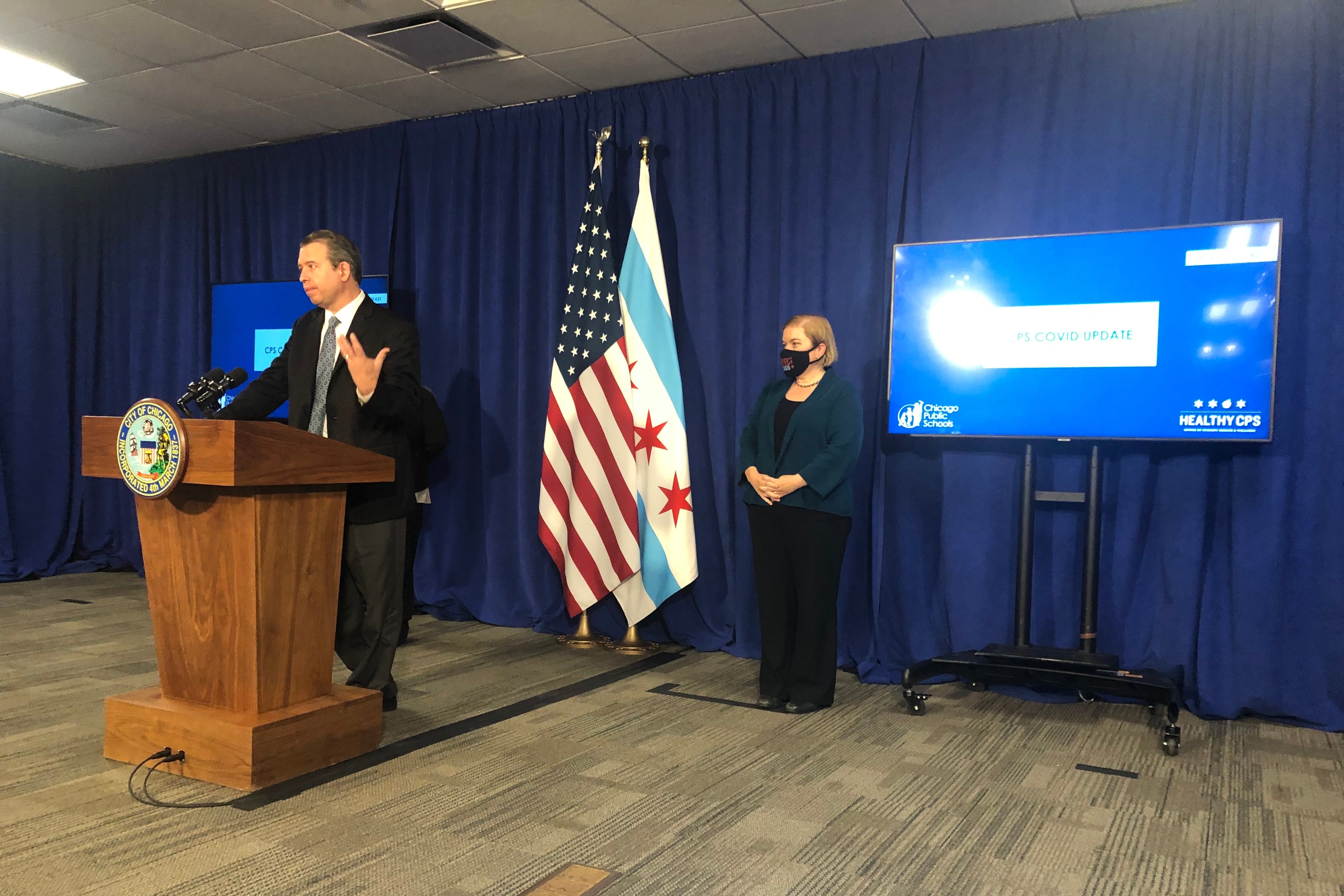 Standing at a podium in a suit and tie, Chicago Public Schools CEO Pedro Martinez explains the new quarantine rules alongside Dr. Allison Arwady, the city’s top health official.