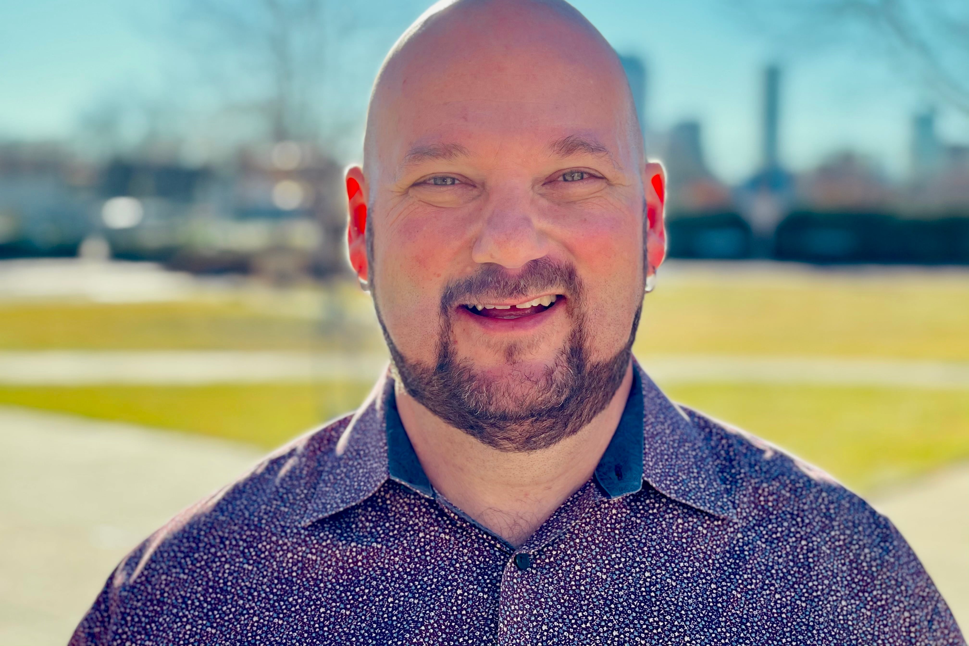A portrait of Denver School Board candidate Scott Esserman. He is wearing a purple shirt and standing in front of a plaza with triangular spaces of lawn with cars parked behind.