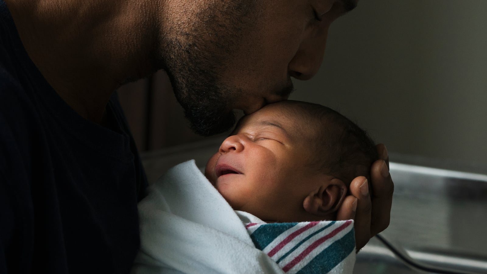 A Black father kissing forehead of newborn son who is wrapped in a baby blanket.