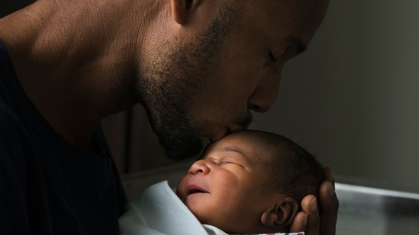 A Black father kissing forehead of newborn son who is wrapped in a baby blanket.