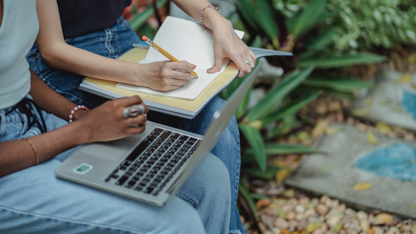 Two female students are seated next to each other on a bench in a park or garden, working on homework together. The student seated to the left is writing something in a notebook with a pencil. The student to the right has a laptop open.