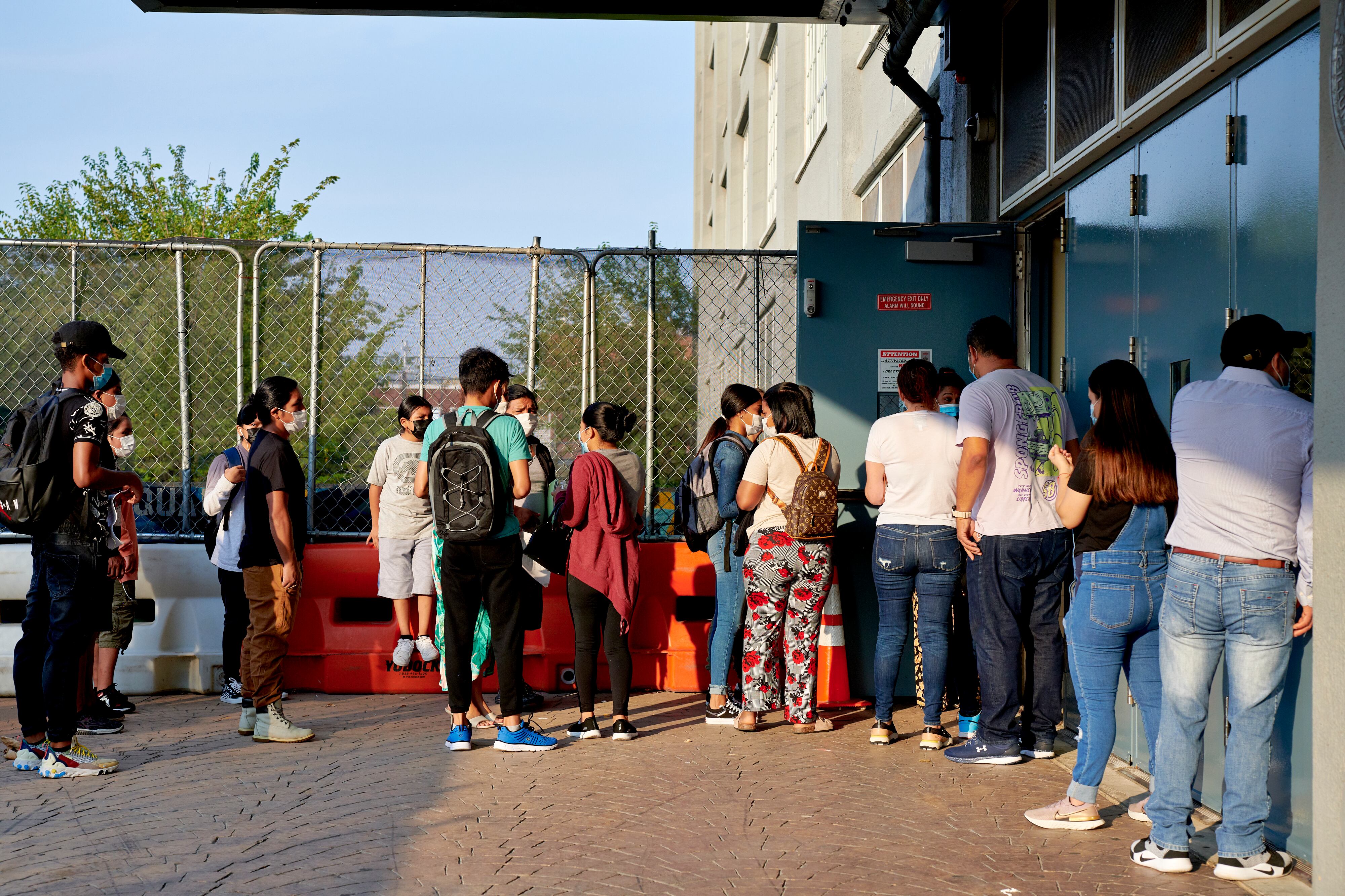 A line of students wait outside a school, with some students along a wall and others along a chain link fence.