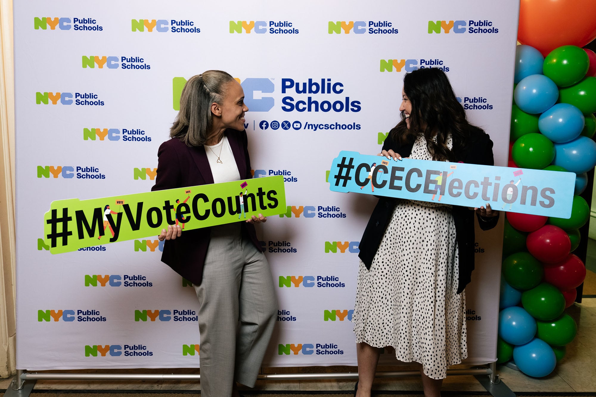 Two women hold colorful signs in front of a backdrop and pose for a photograph.