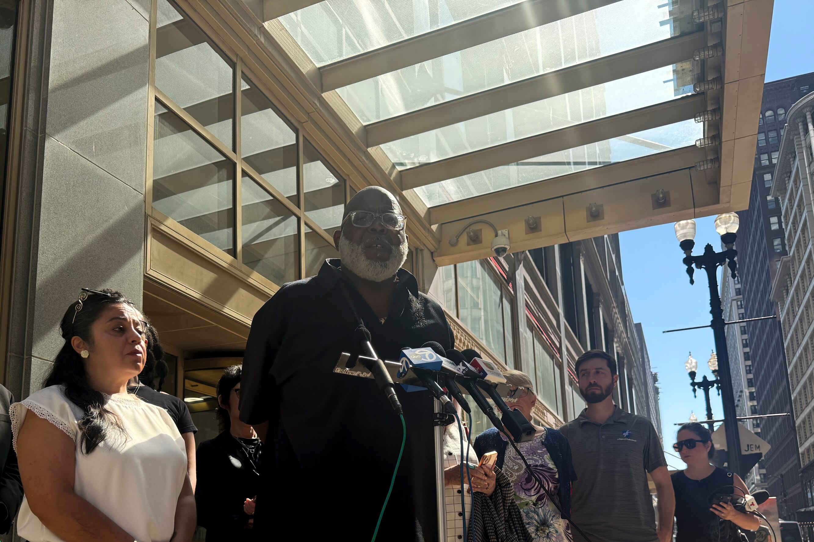 A Black male standing on a sidewalk, speaking into the microphones.