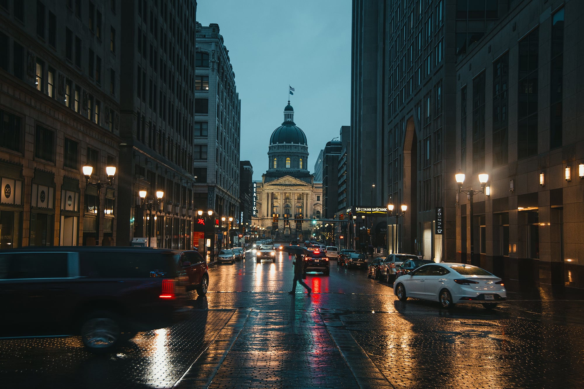A photograph of the Indiana Statehouse after the sun has set with a person holding an umbrella crosses the street while cars zoom around.