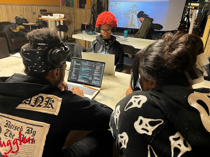 A photograph of the backs of a Black male and a Black female, both looking at the laptop on the table in front of them. There are lines of code on the laptop's screen.
