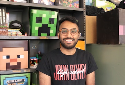 A man with short dark hair, wearing glasses and wearing a black shirt sits in front of a bookshelf with Minecraft memorabilia.