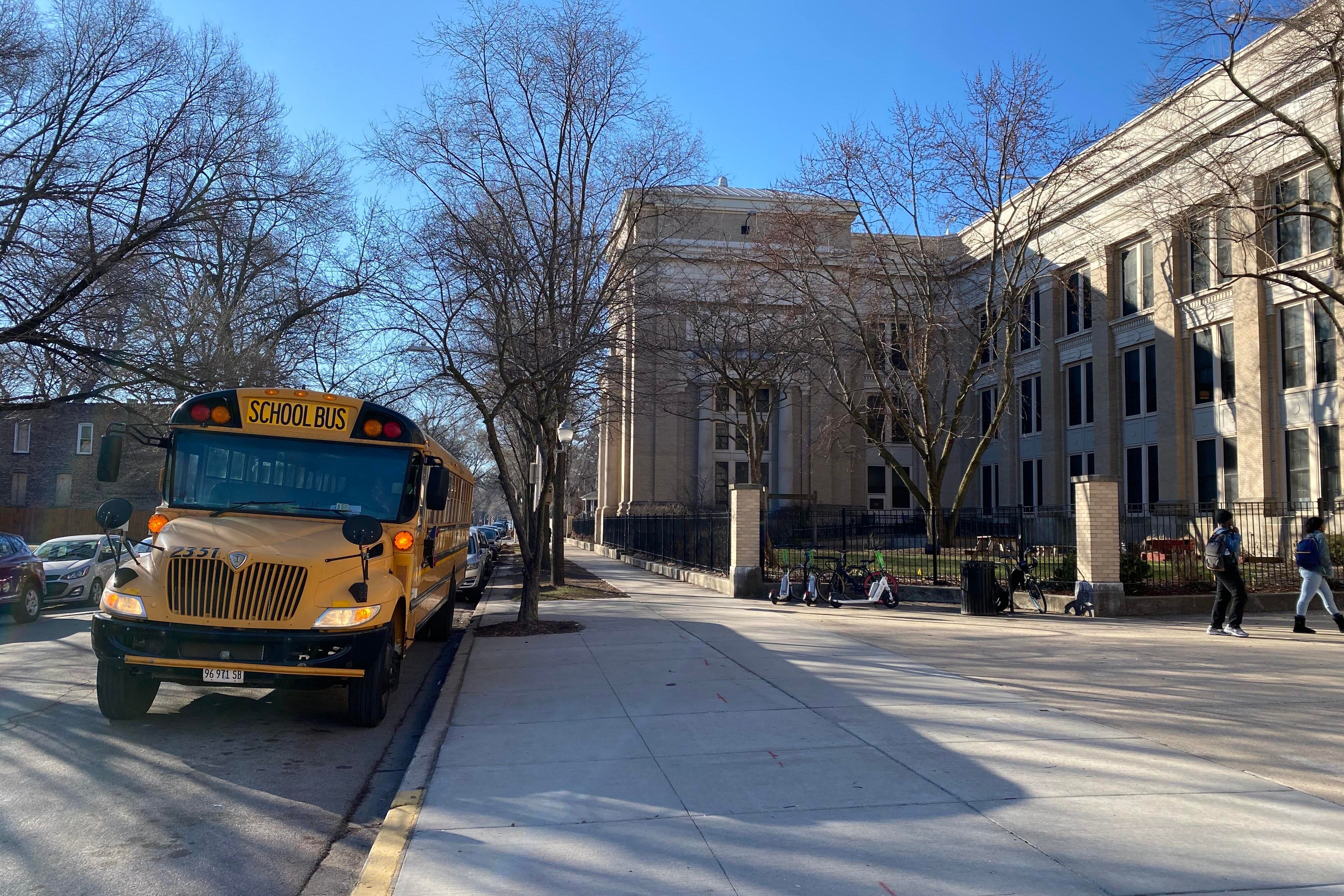A yellow school bus is parked outside of a large stone building with a large sidewalk in the foreground and blue sky in the background.