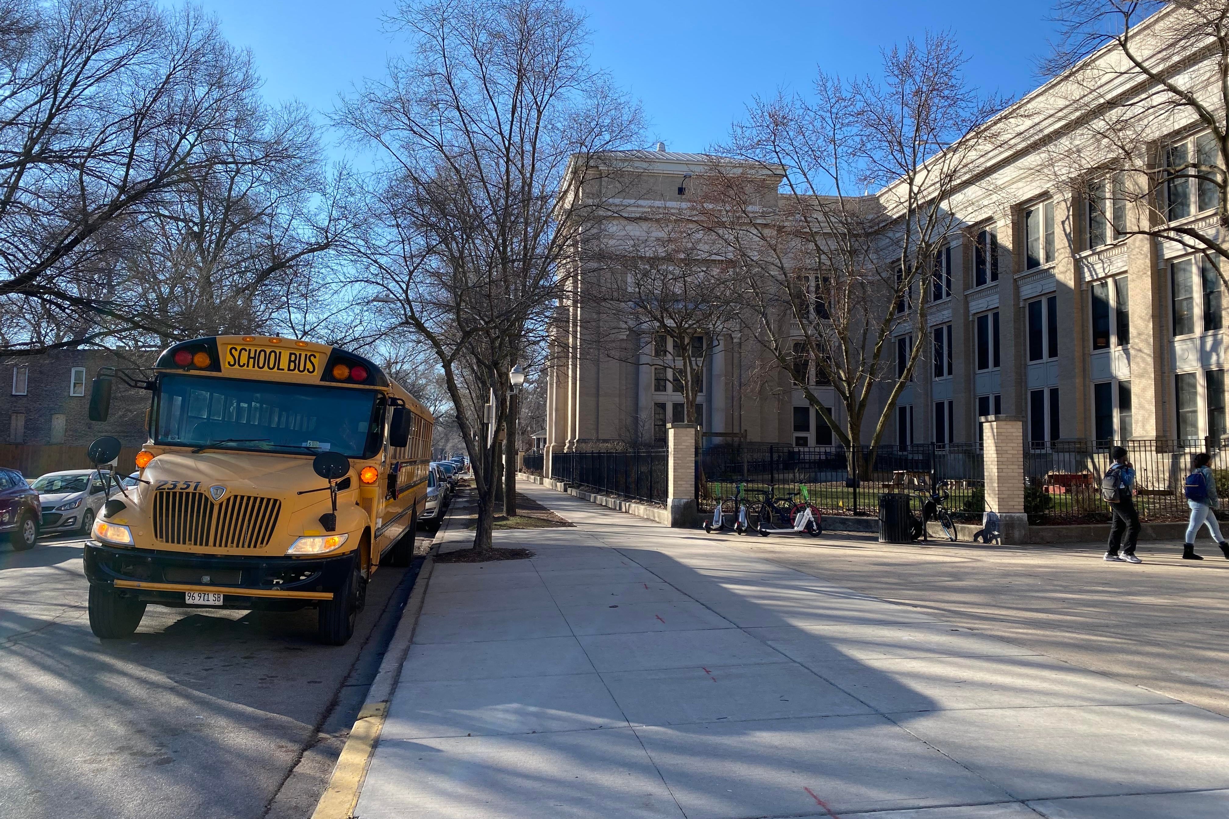 A yellow school bus is parked outside of a large stone building with a large sidewalk in the foreground and blue sky in the background.