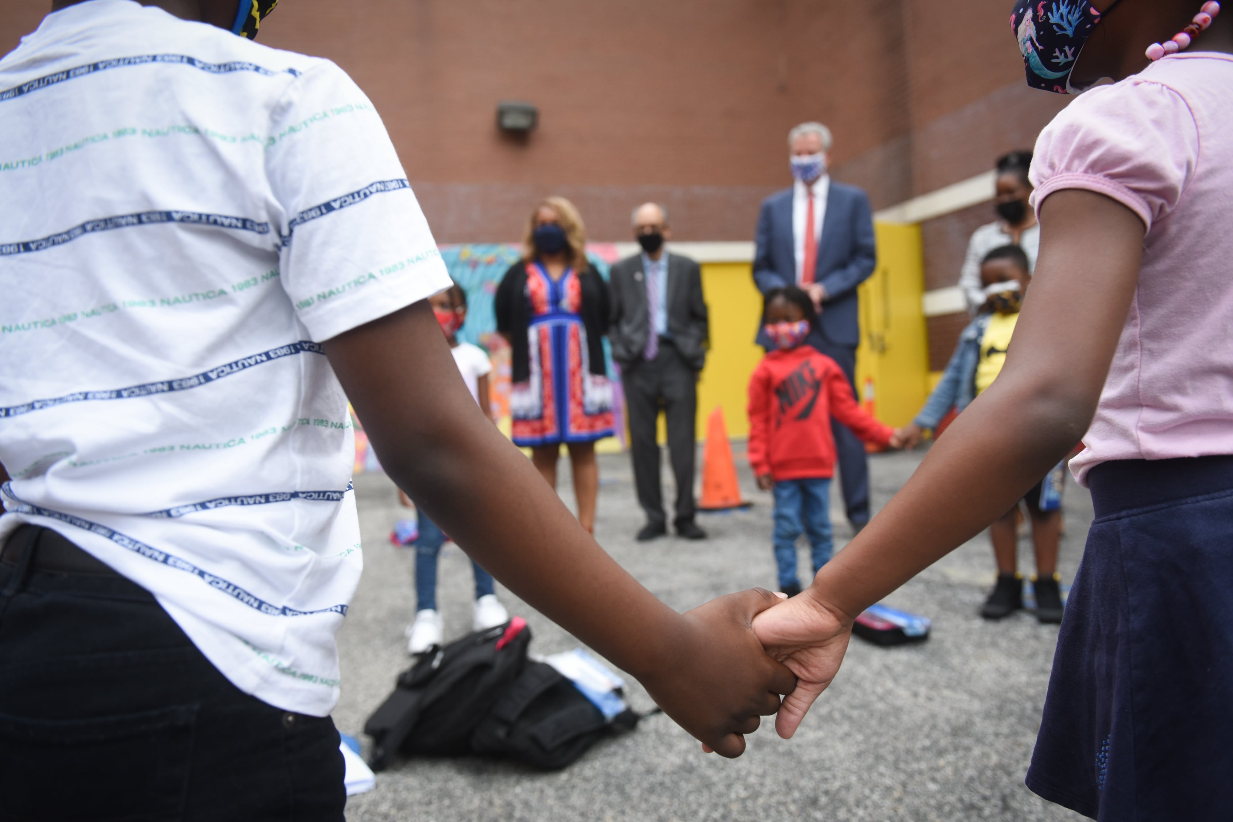 Two young students hold hands in the foreground while a group of adults and students stand in the background.