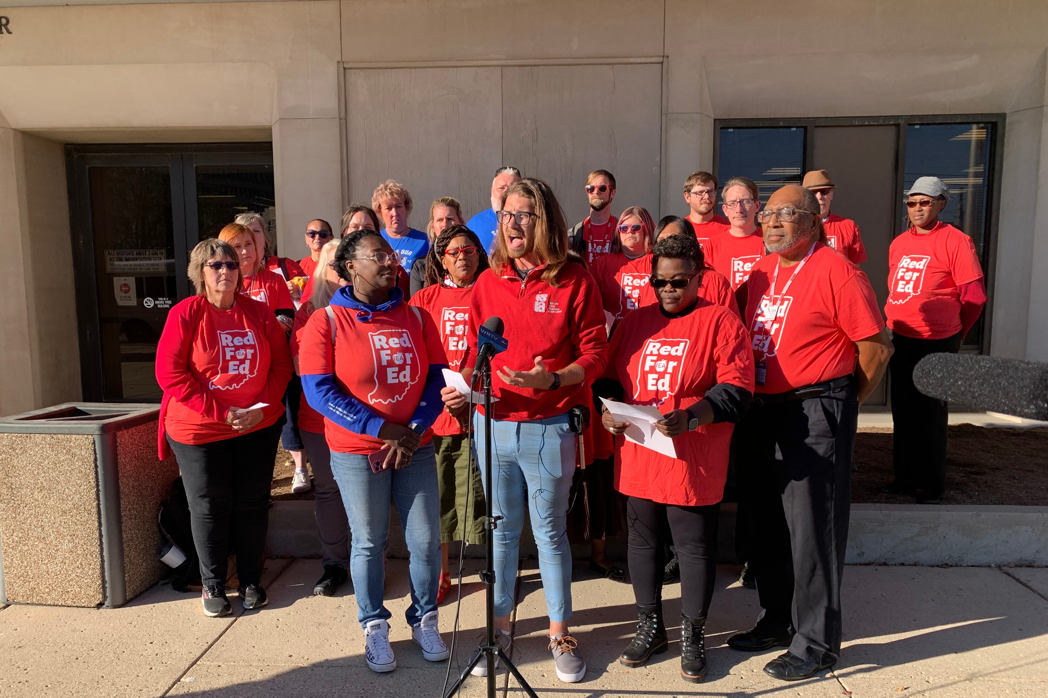 A man in a red shirt stands in front of a group of about a dozen other people in red shirts and speaks into a microphone.