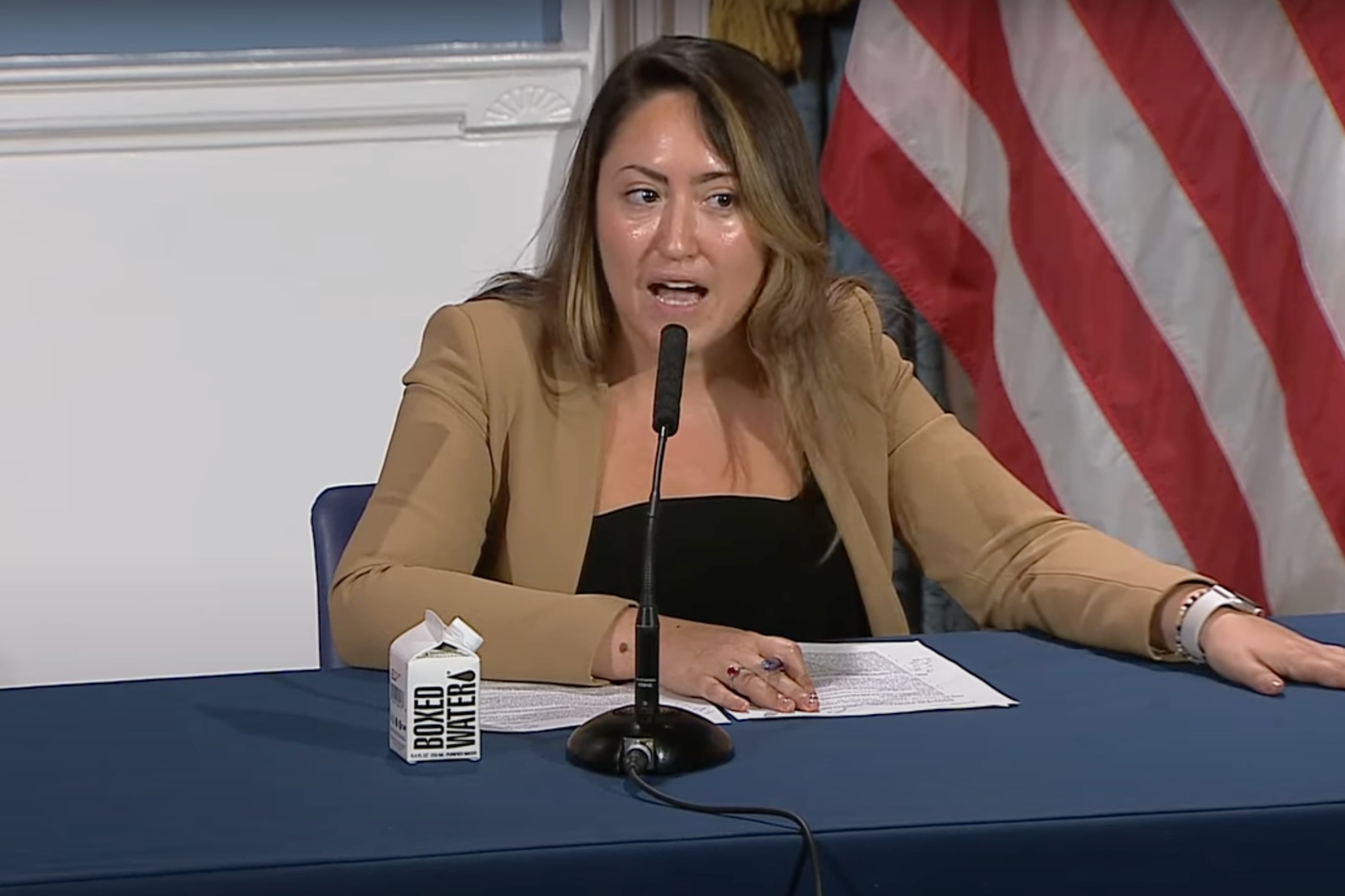 A woman in a tan blazer sits at a blue table with an American flag behind her.
