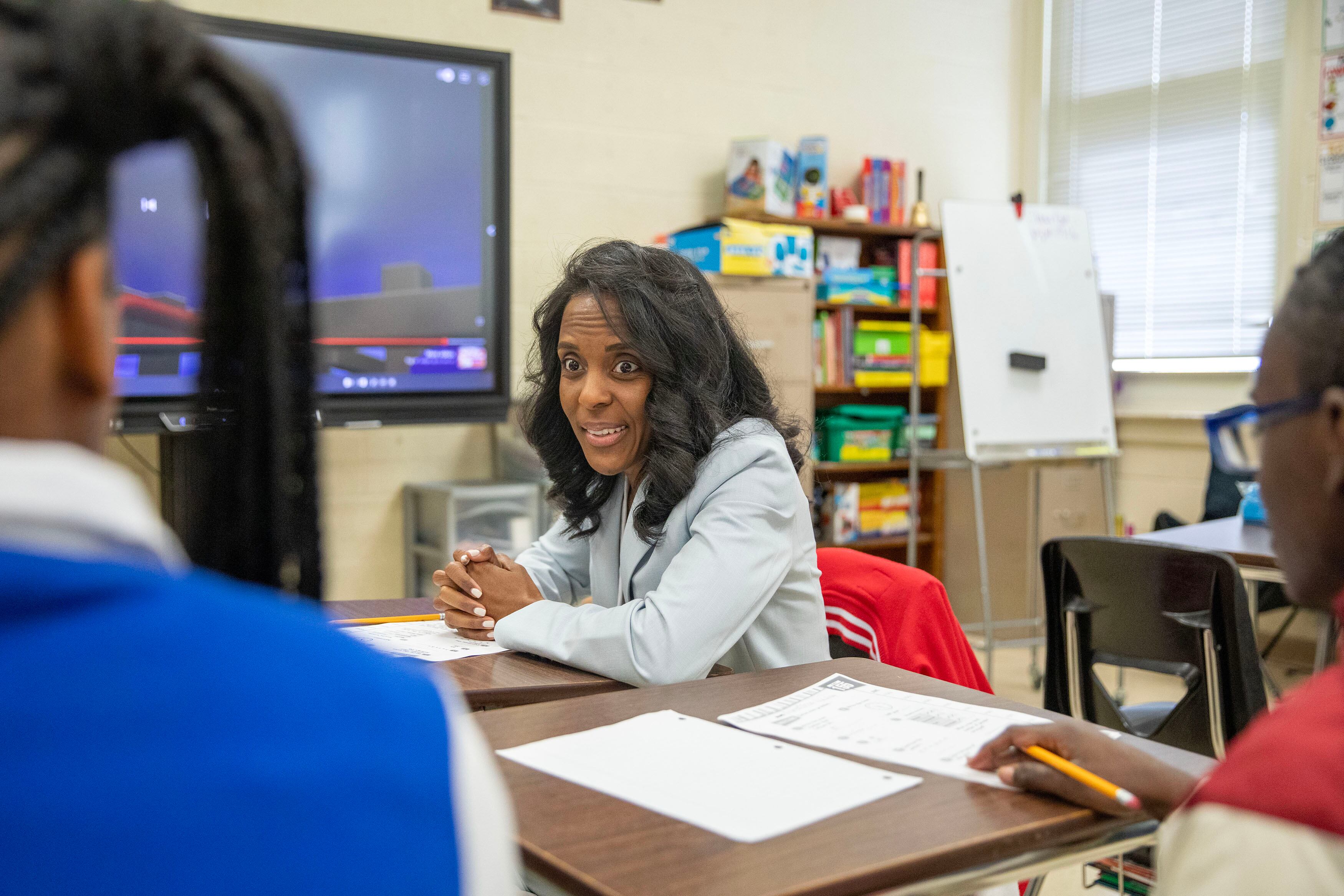 An adult woman wearing a light suit sits at a wooden desk facing young students with their backs toward the camera in a classroom.