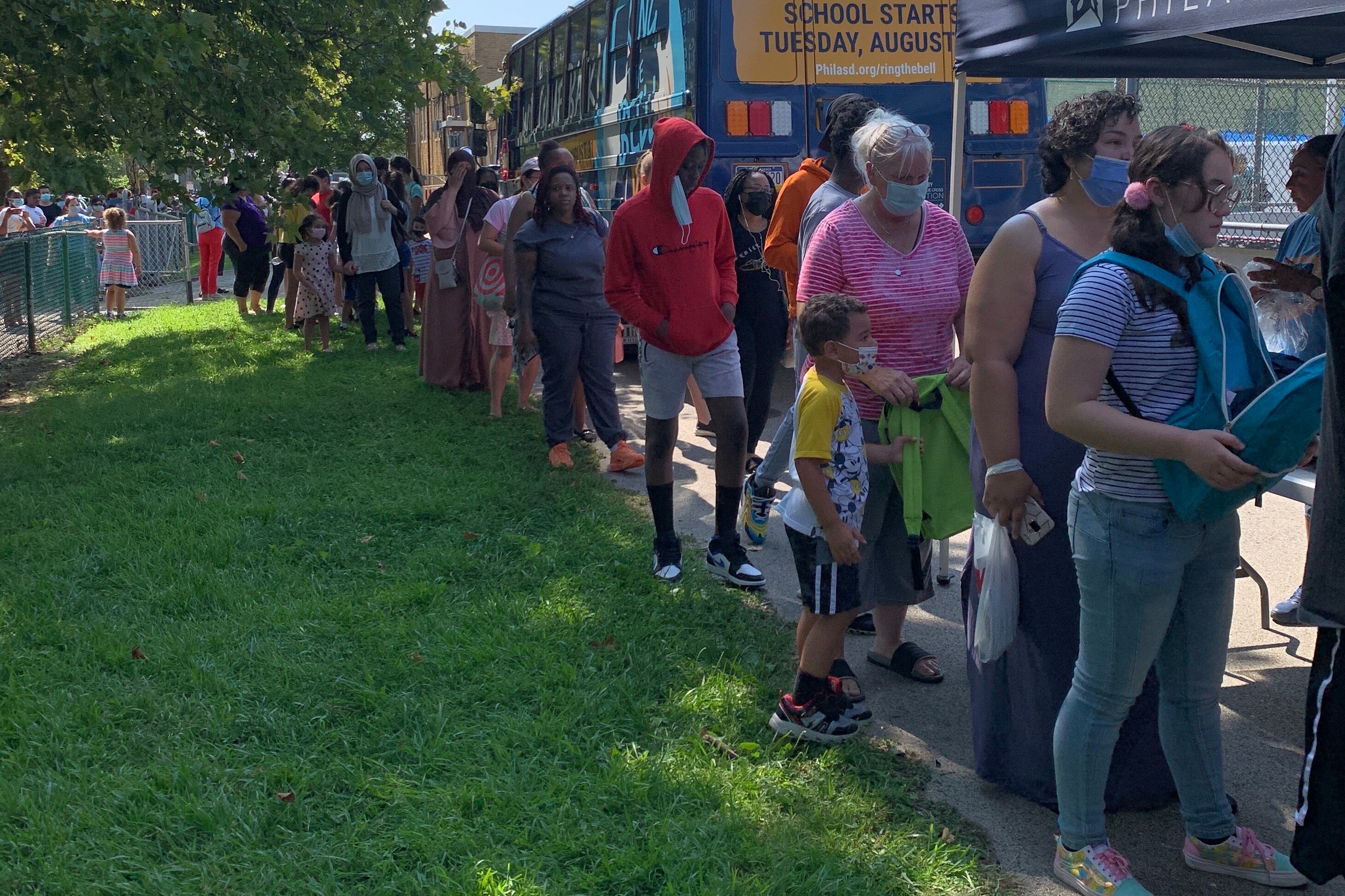 A long line of families wait near Cottman Avenue in the Northeast. They stand on a sidewalk next to a grassy area.