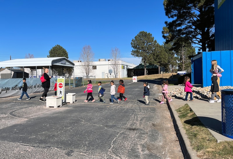 A group of young students cross a street in a line with a teacher on each end of the line. Trees and buildings are in the background.