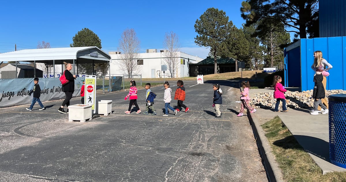 A group of young students cross a street in a line with a teacher on each end of the line. Trees and buildings are in the background.