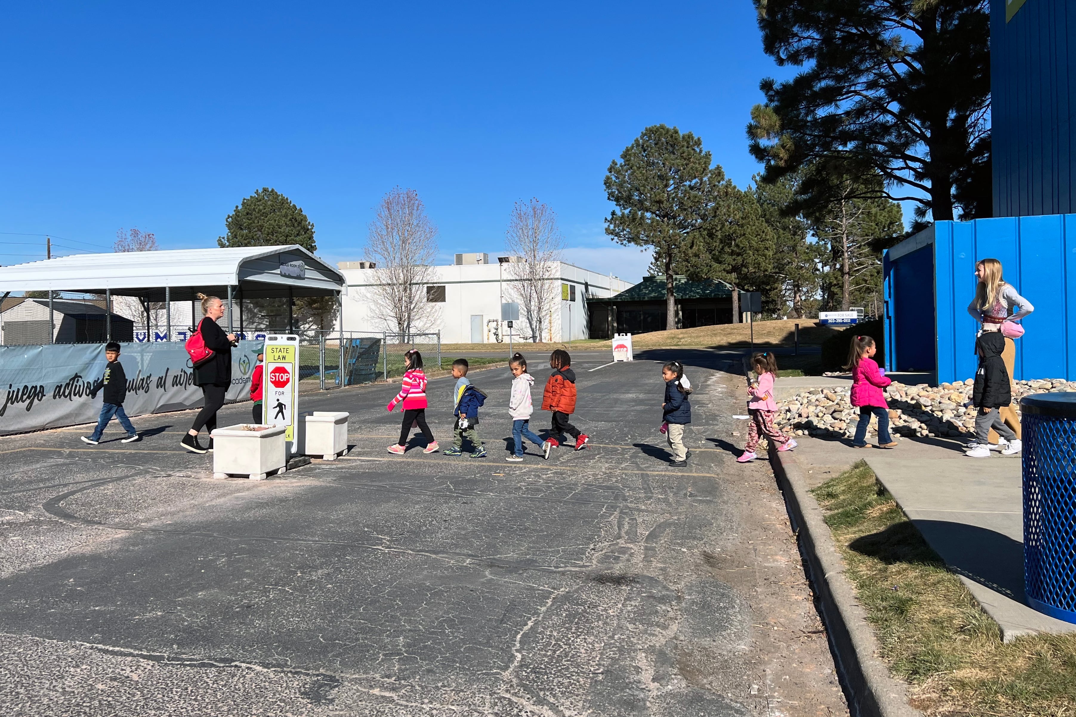 A group of young students cross a street in a line with a teacher on each end of the line. Trees and buildings are in the background.