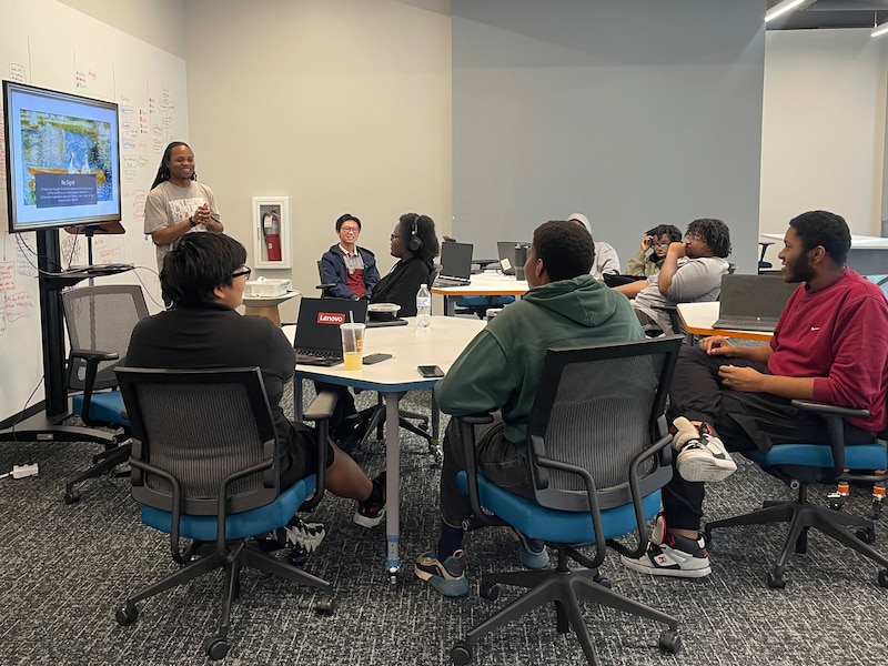 A class of young adults sit at their desks while an adult stands at the side of the class in front of a TV screen.